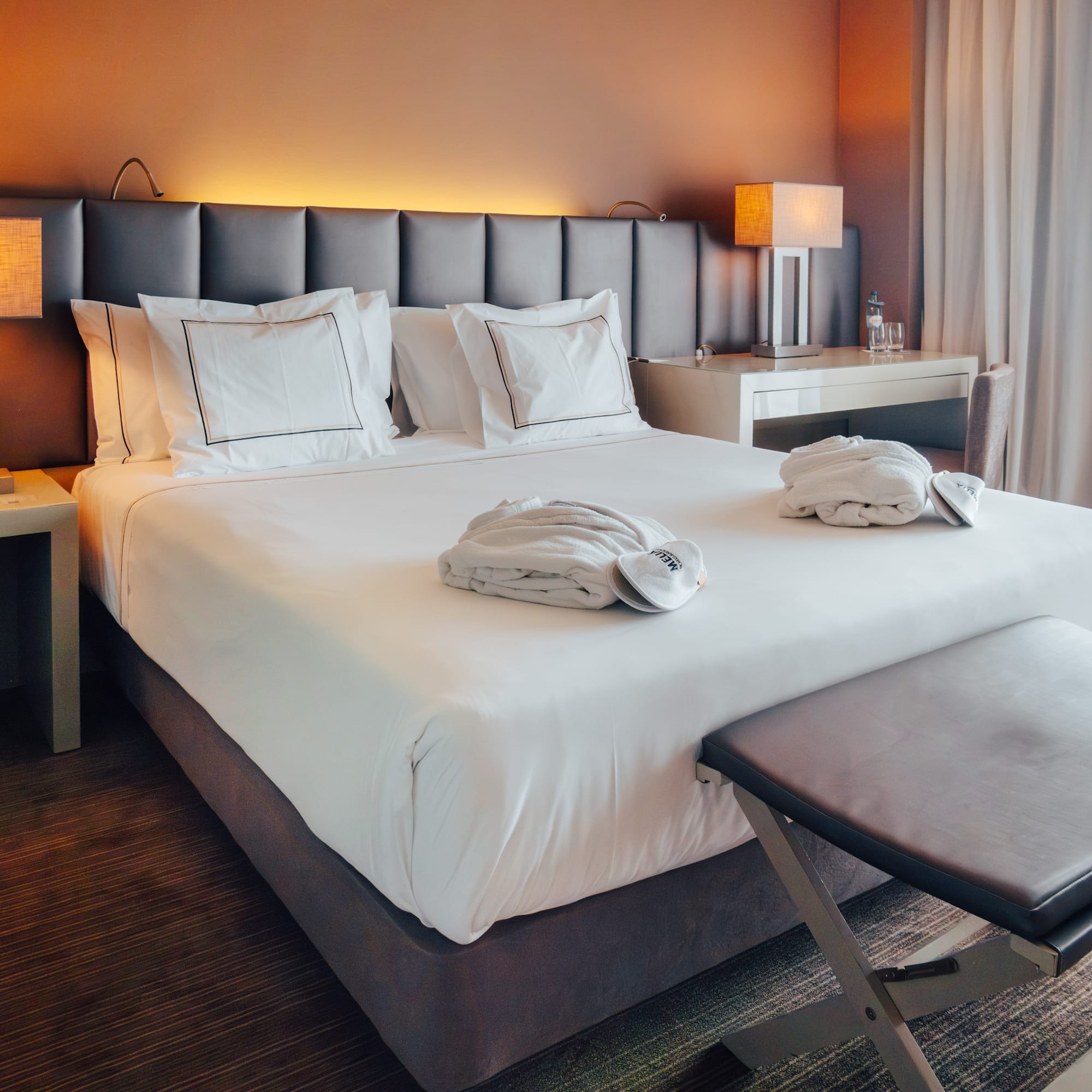 Modern hotel room with bed, white linens, bathrobes, lamps, and sheer curtains.