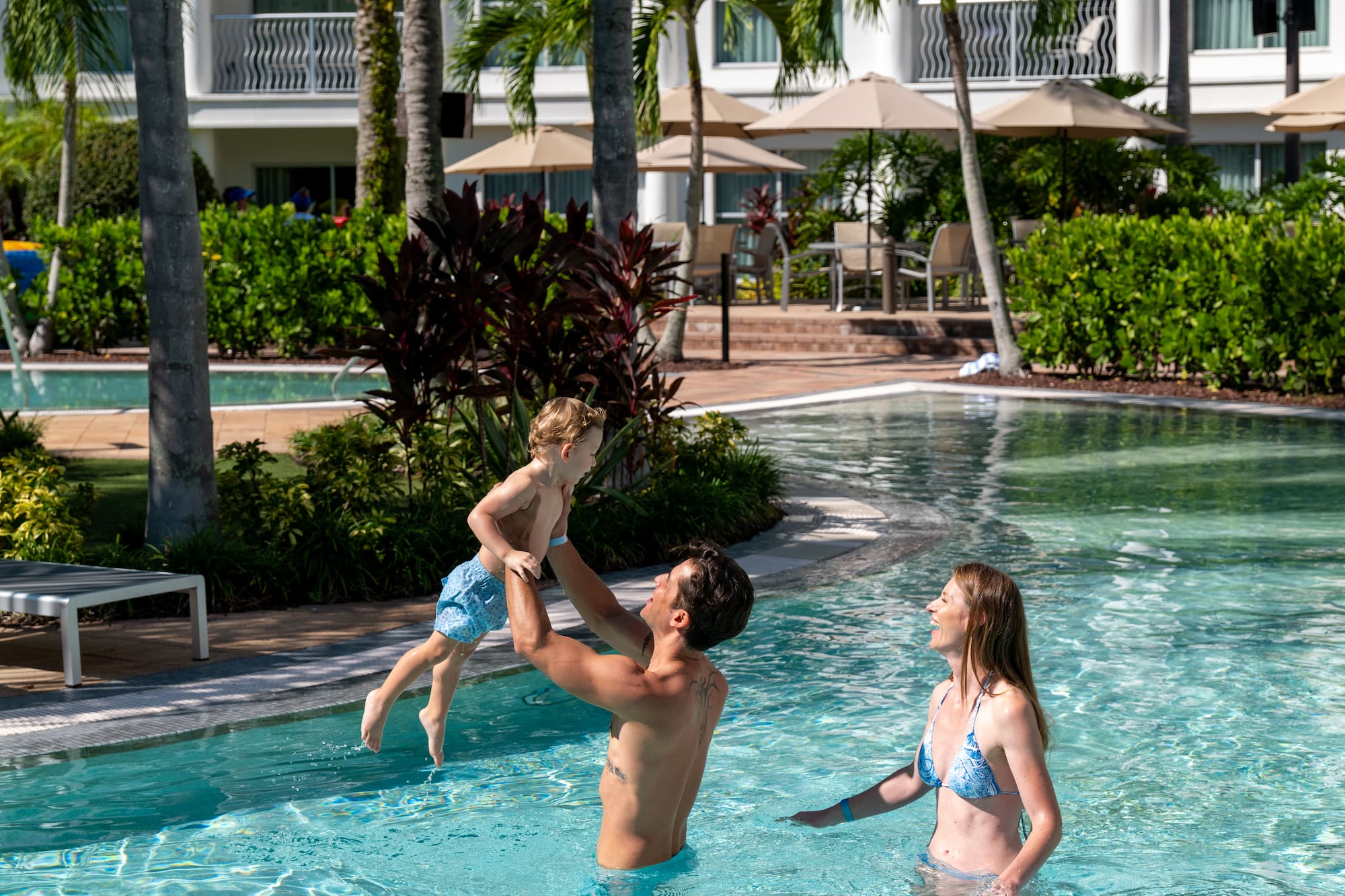 Dad lifting child in resort swimming pool with mom, palm trees, and hotel.