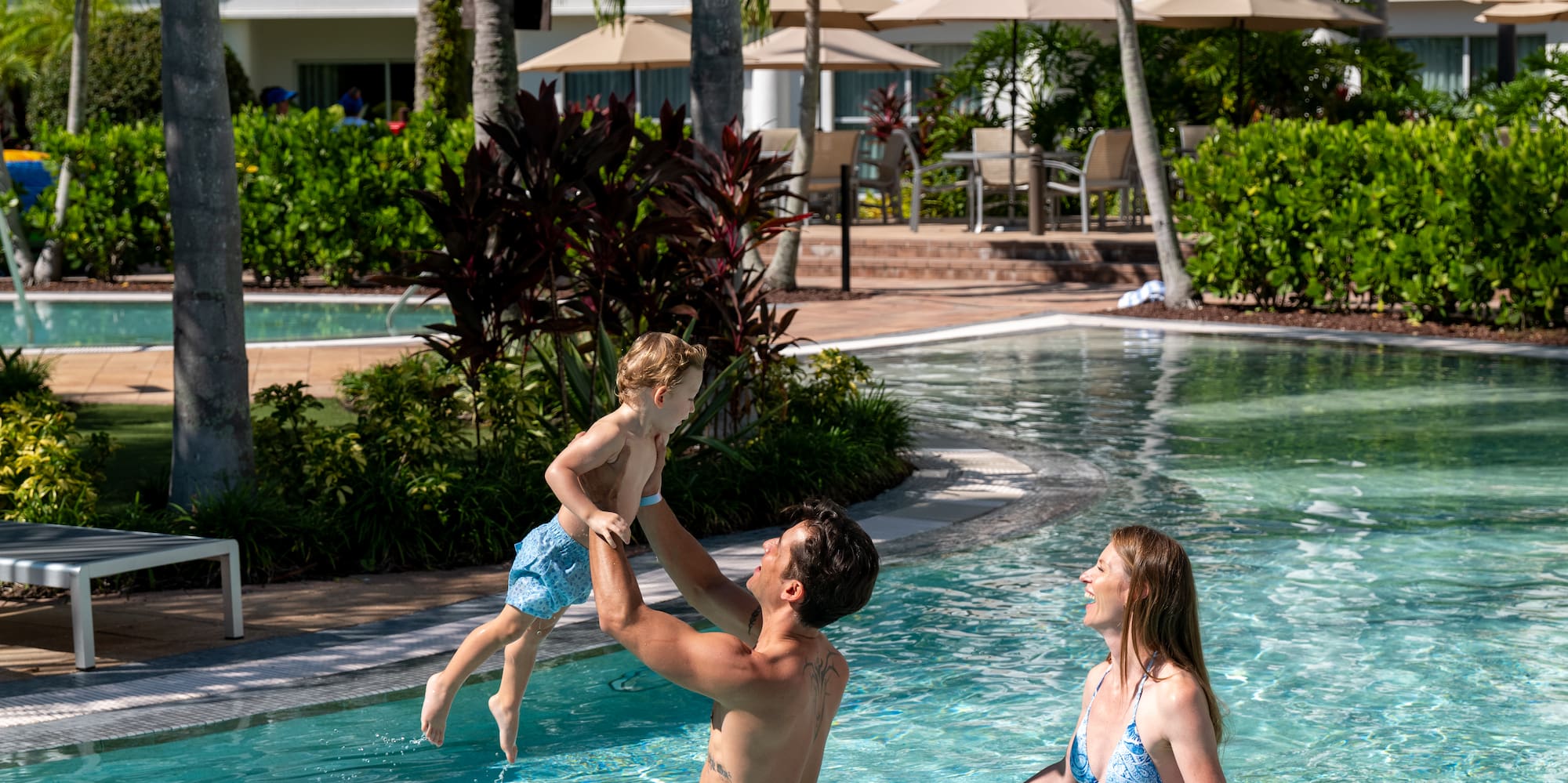 Dad lifting child in resort swimming pool with mom, palm trees, and hotel.