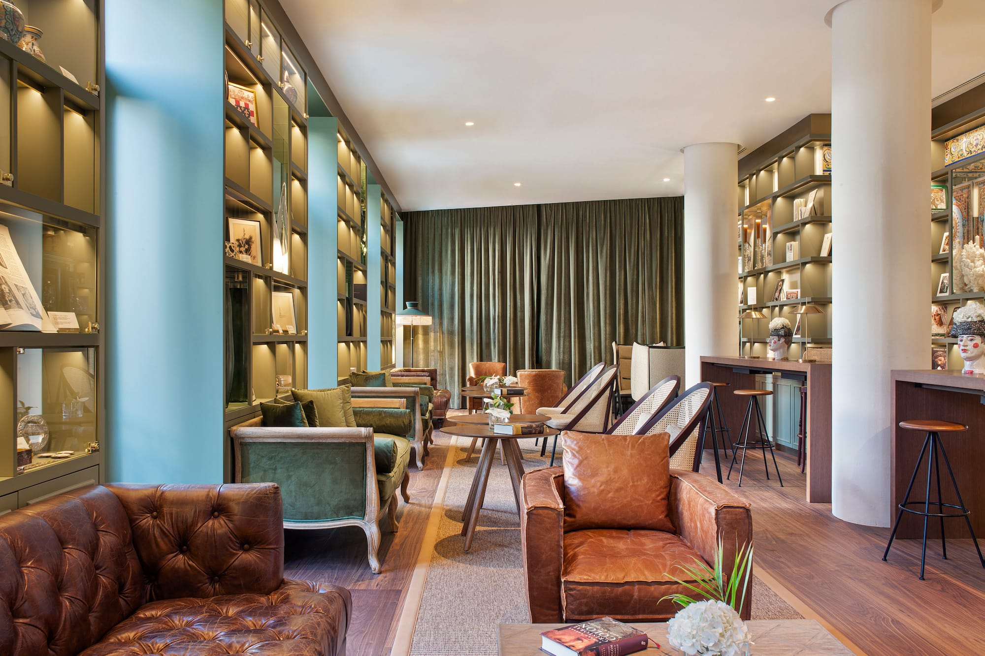 Hotel lounge with brown leather and green velvet seating, library shelves, bar.