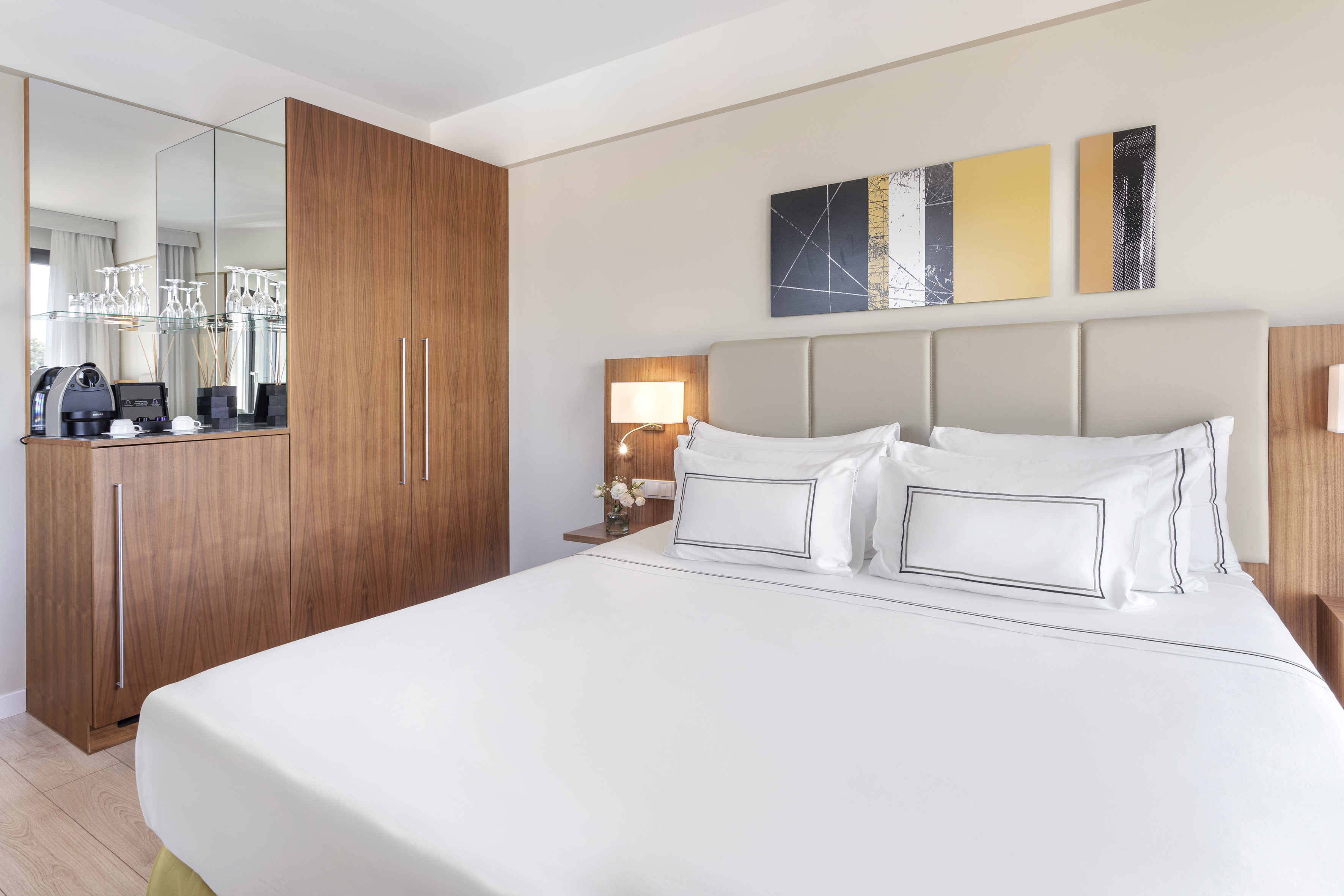 Modern hotel room with white bed, abstract art, and coffee bar.