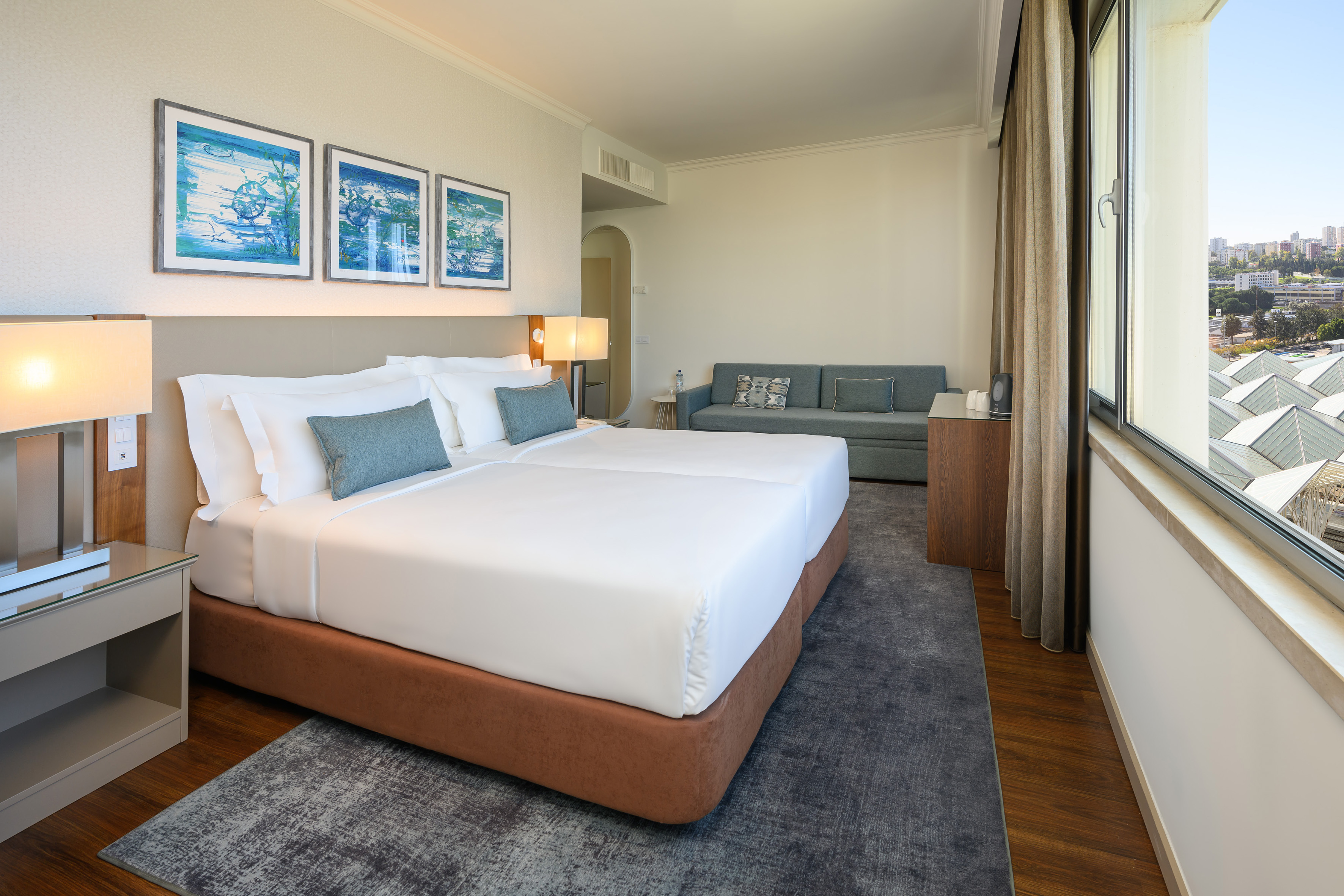 Modern hotel room with twin beds, grey sofa, and city view from window.