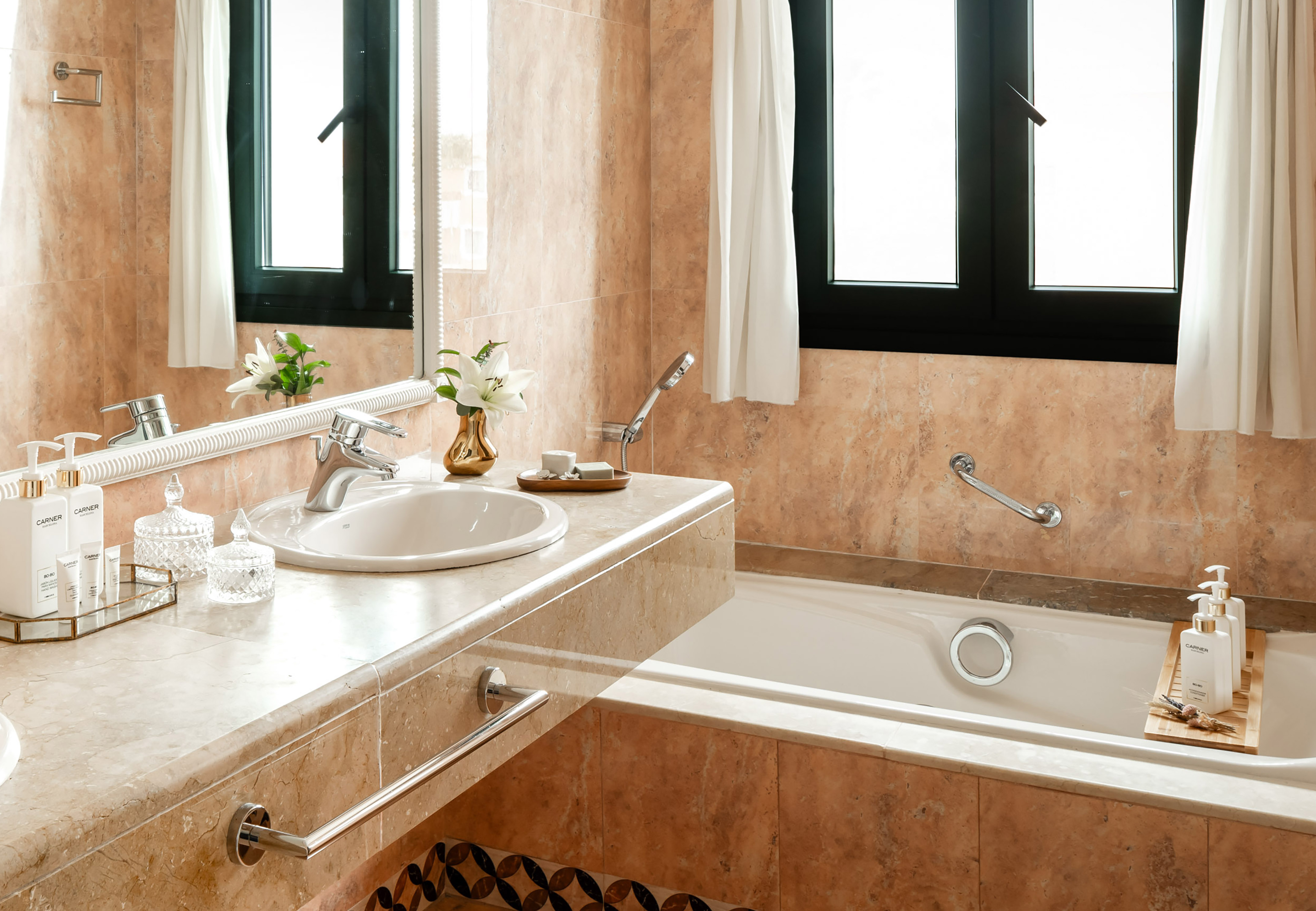 Warm marble bathroom with bathtub, sink vanity, mirror, and natural light.