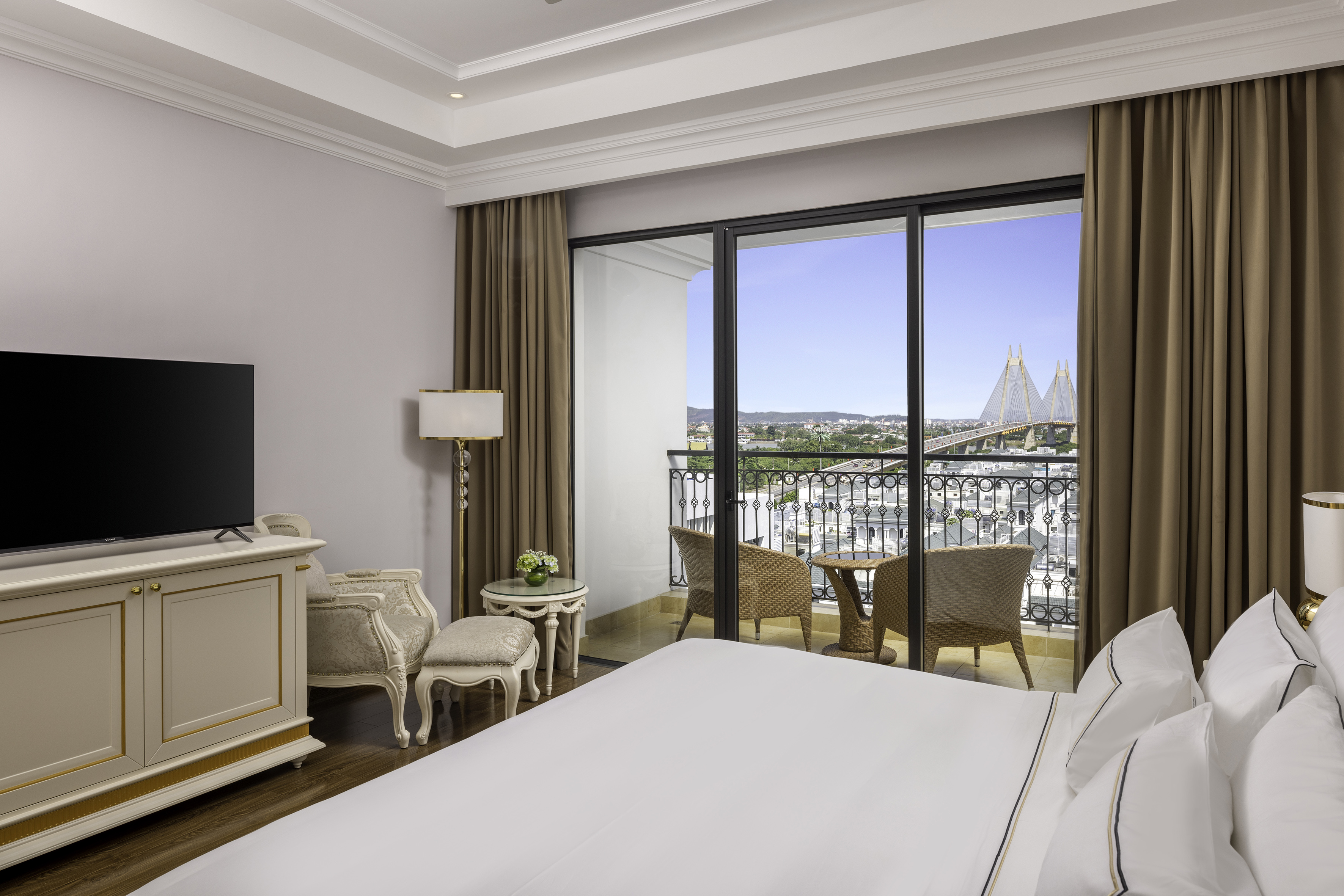Elegant hotel bedroom with balcony, city view, and suspension bridge.