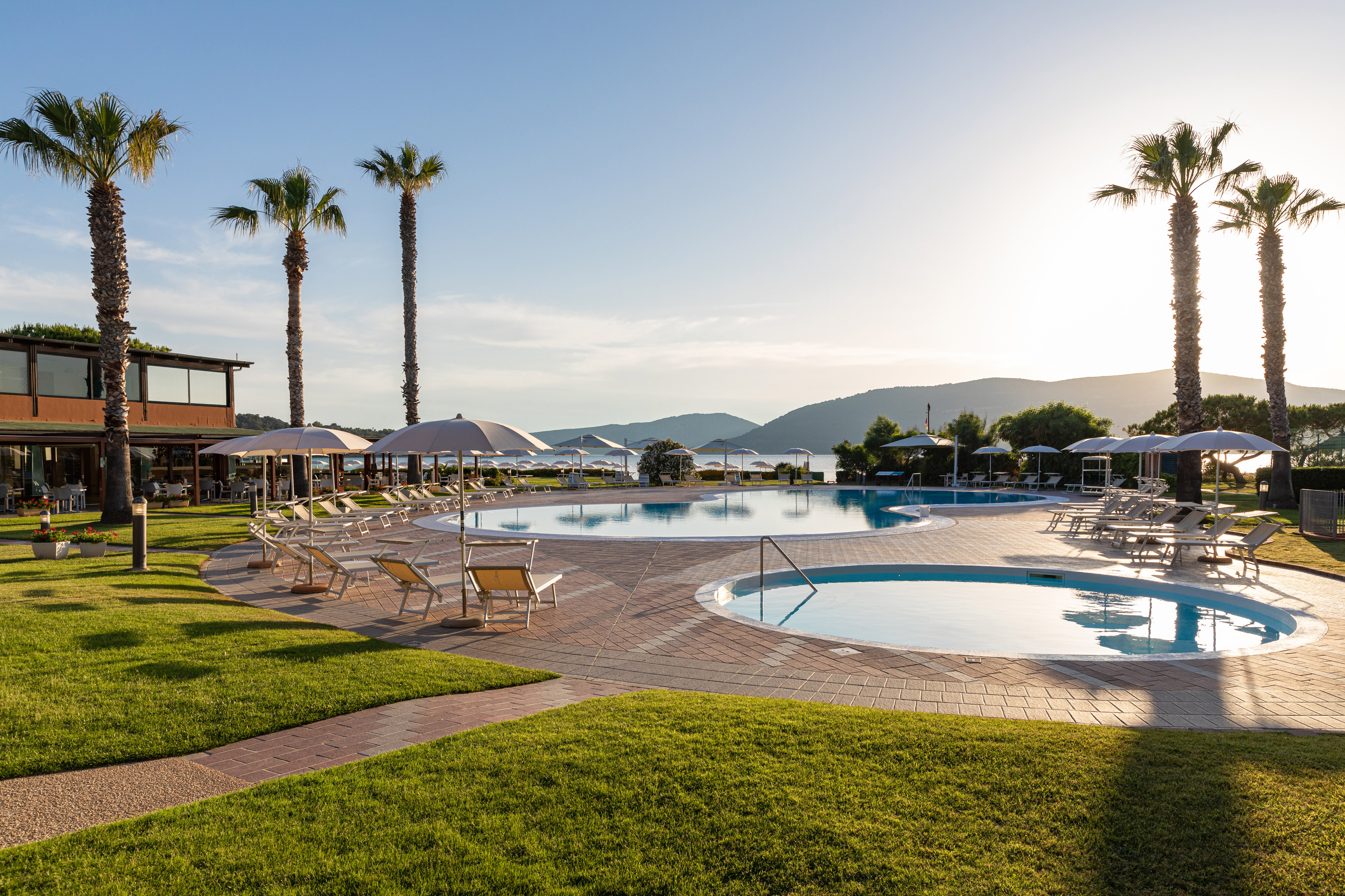 Sunset at resort pools with palm trees, loungers, grass, and a bay view.