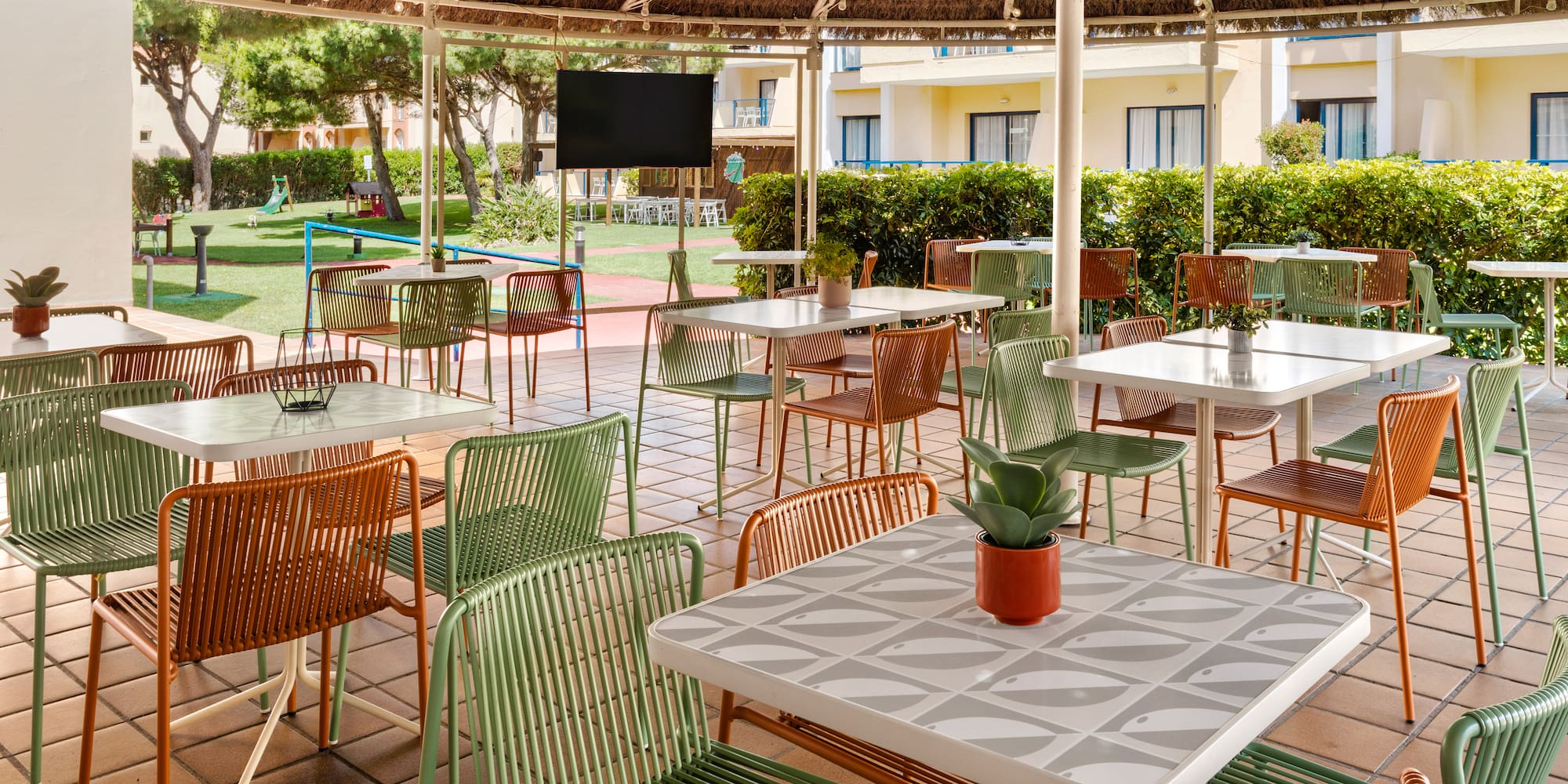 Hotel outdoor patio dining with green/orange chairs, patterned tables, thatched roof.