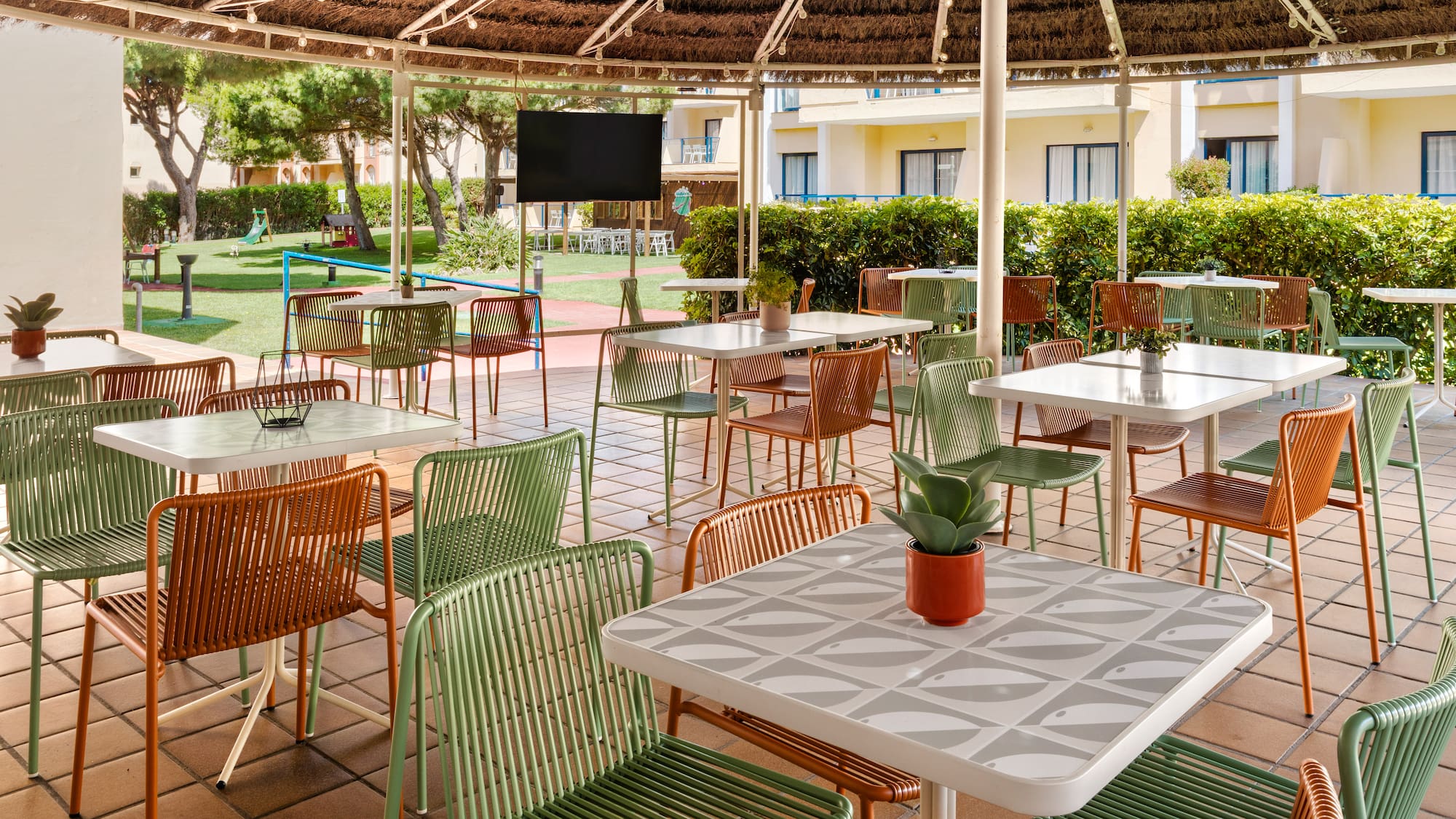 Hotel outdoor patio dining with green/orange chairs, patterned tables, thatched roof.
