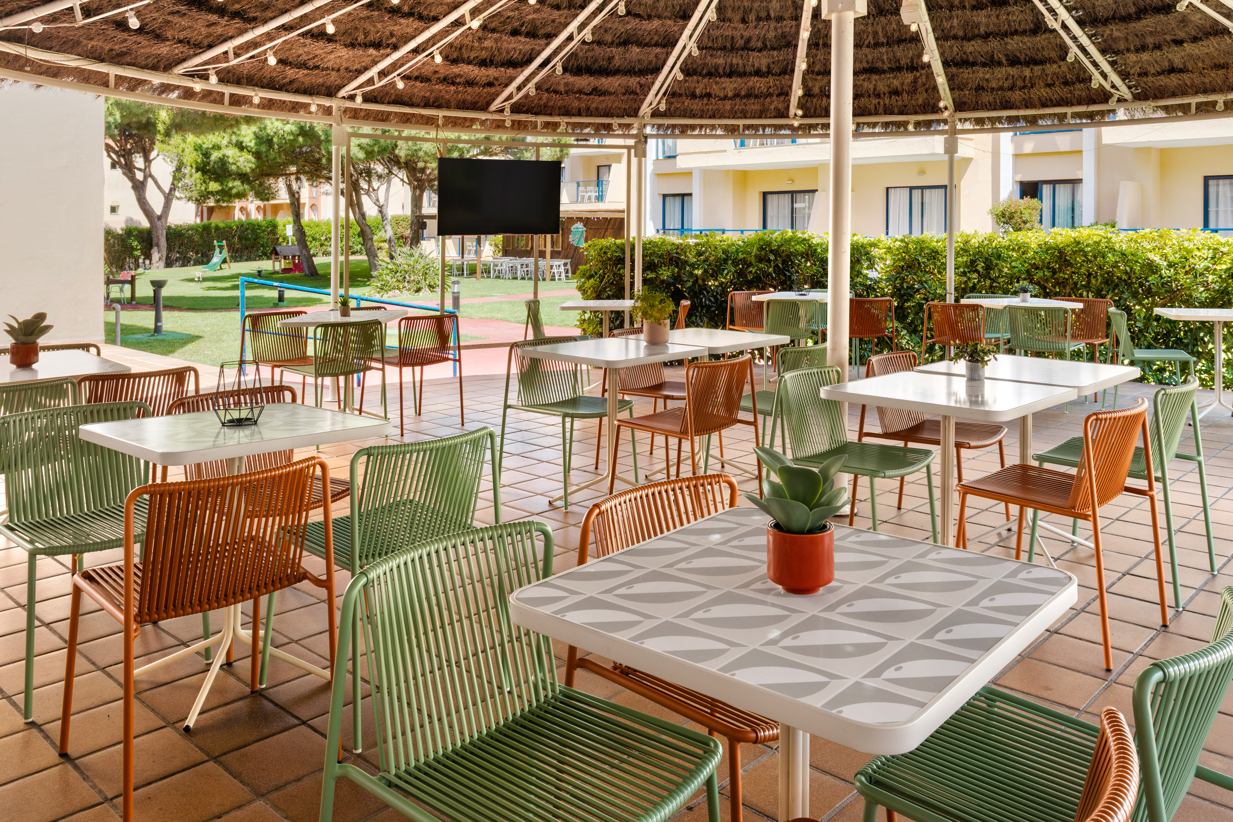 Hotel outdoor patio dining with green/orange chairs, patterned tables, thatched roof.