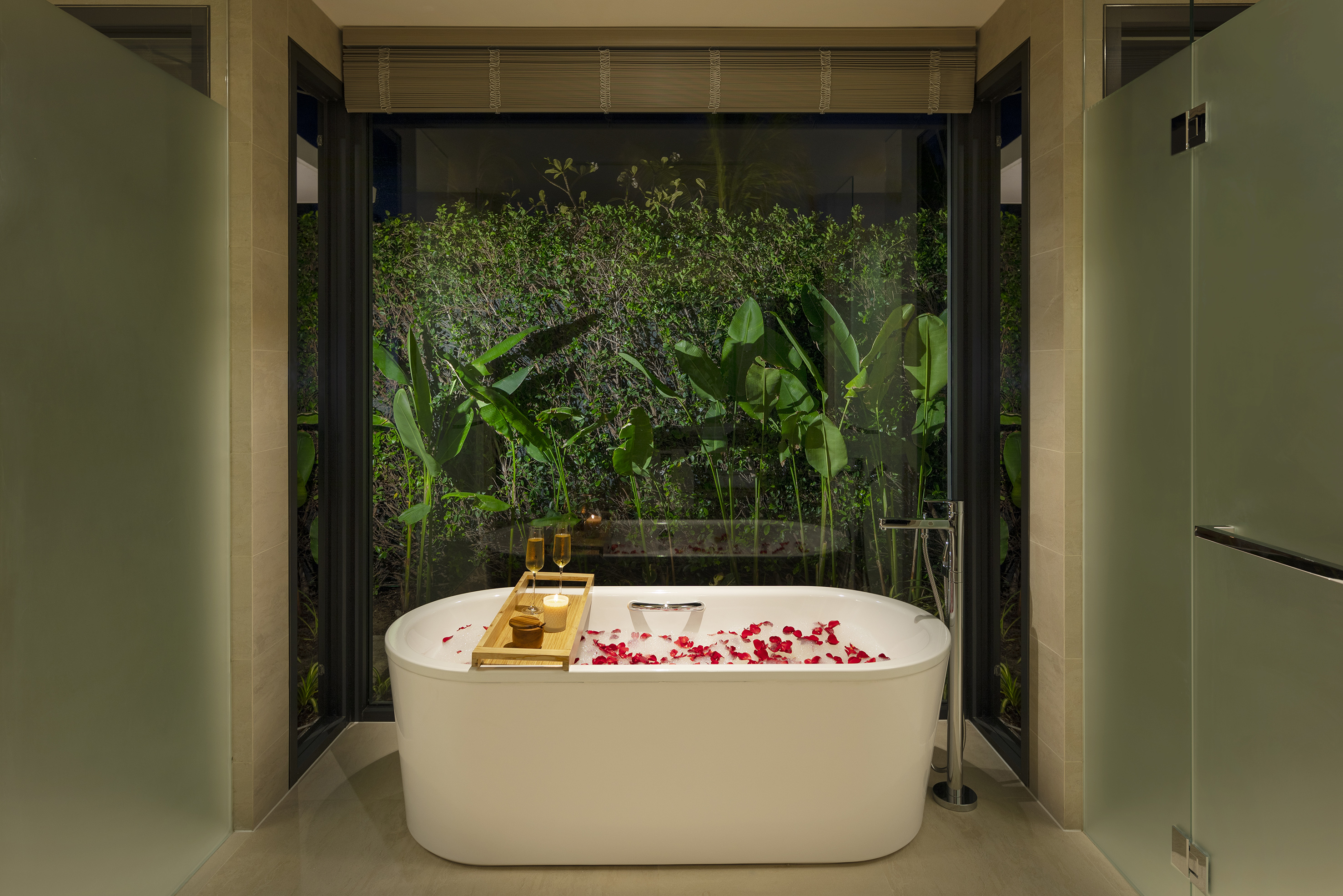 Luxury bathtub with rose petals, champagne, and lush garden view.
