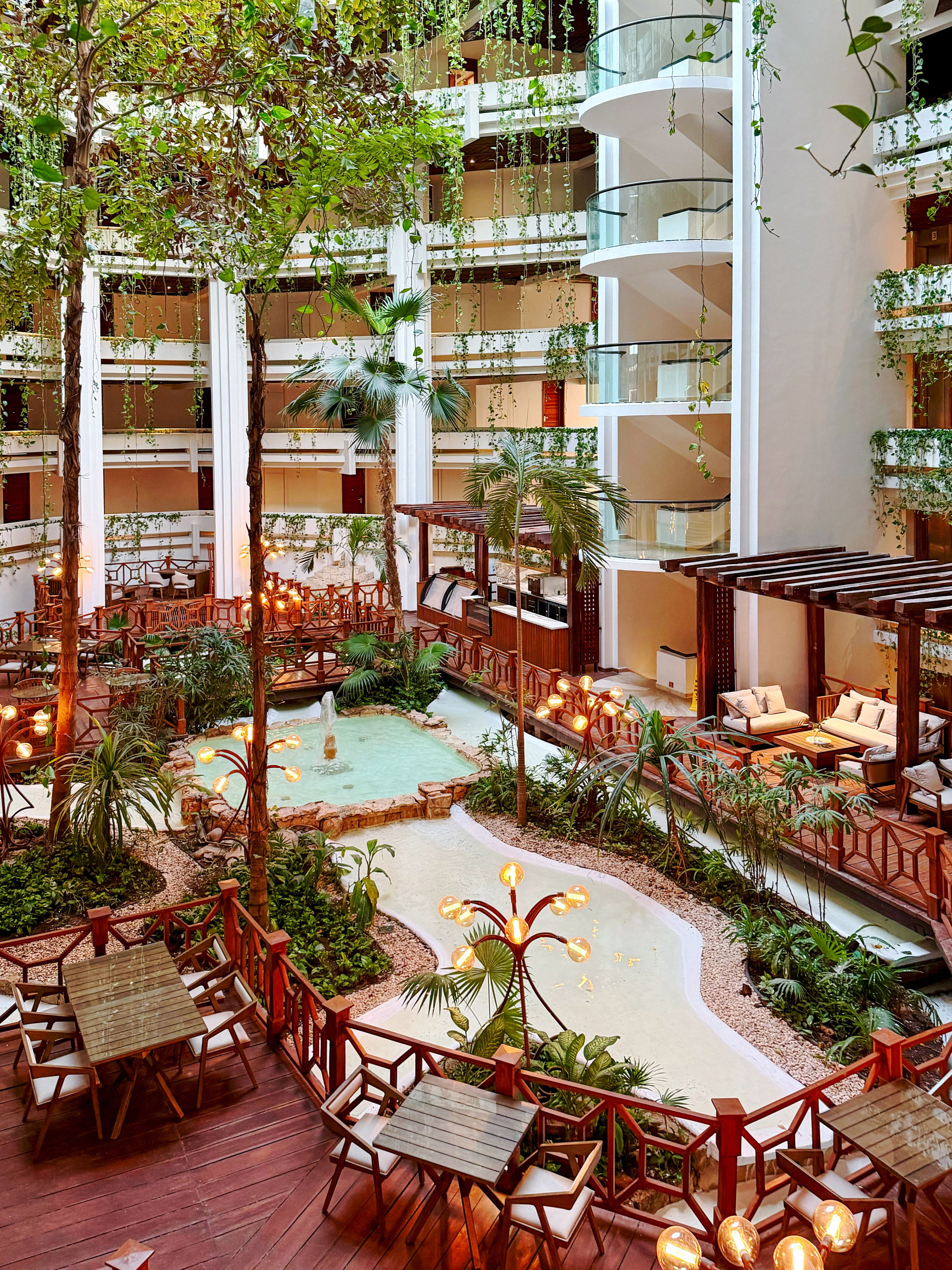 Lush tropical resort atrium with indoor garden, water feature, seating, and warm lighting.