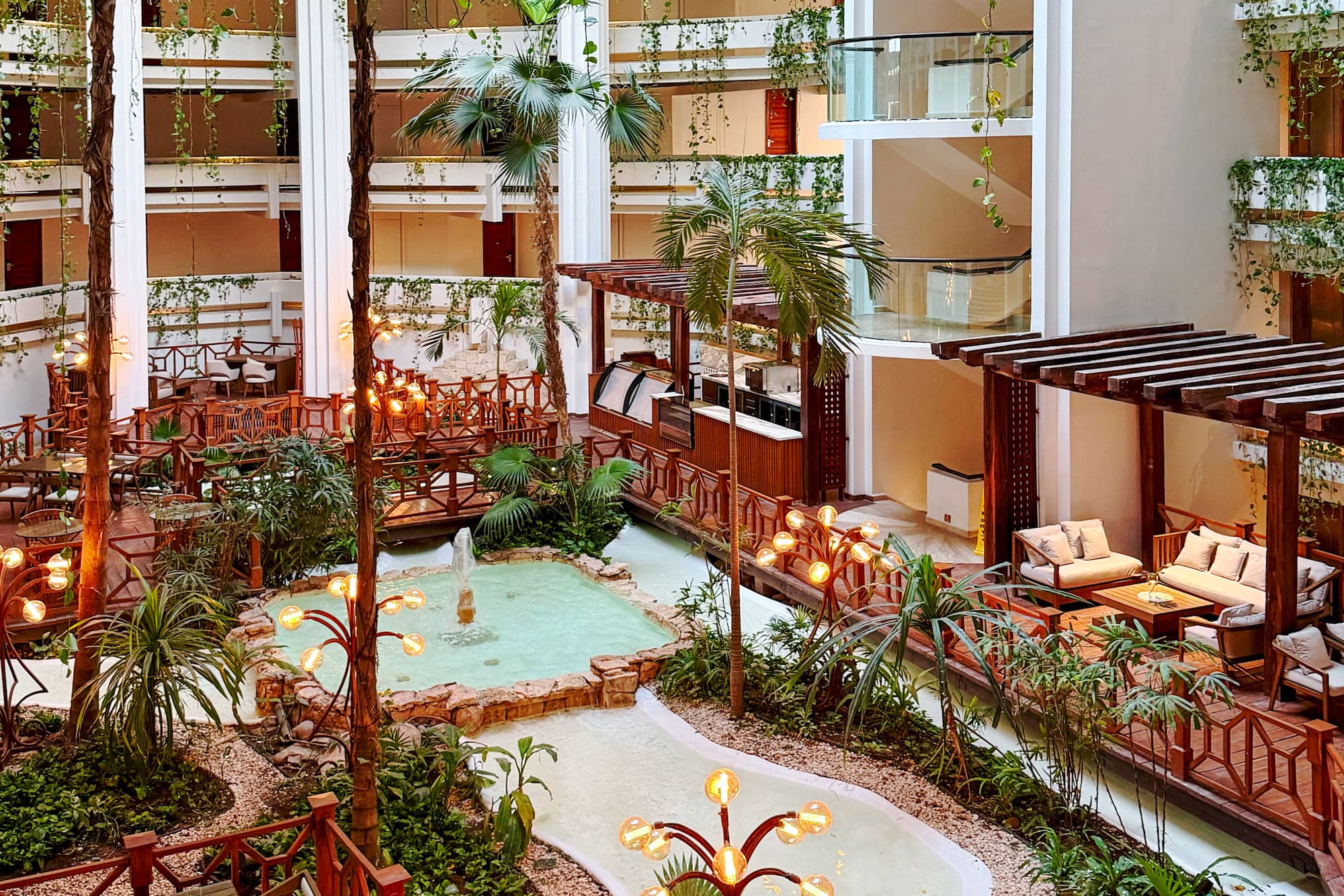 Lush tropical resort atrium with indoor garden, water feature, seating, and warm lighting.