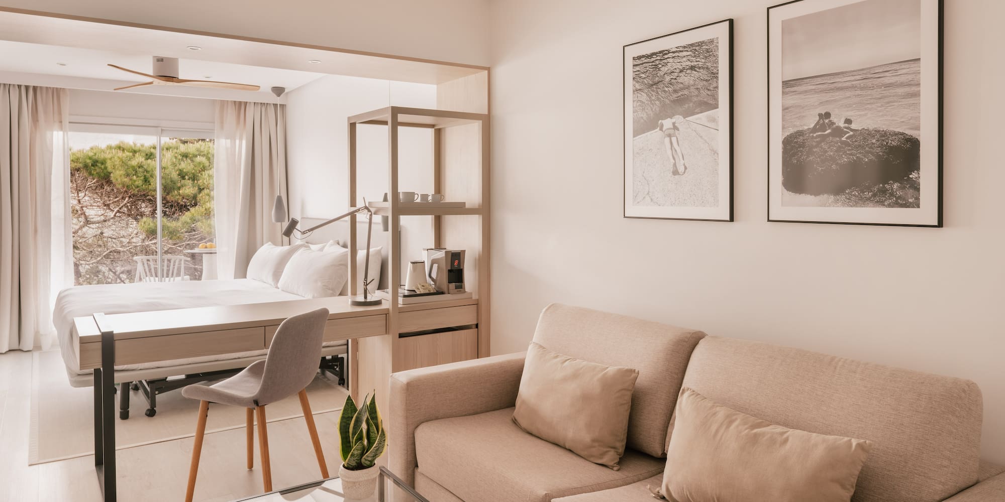 Modern hotel suite with beige sofa, desk, bed, and large window.