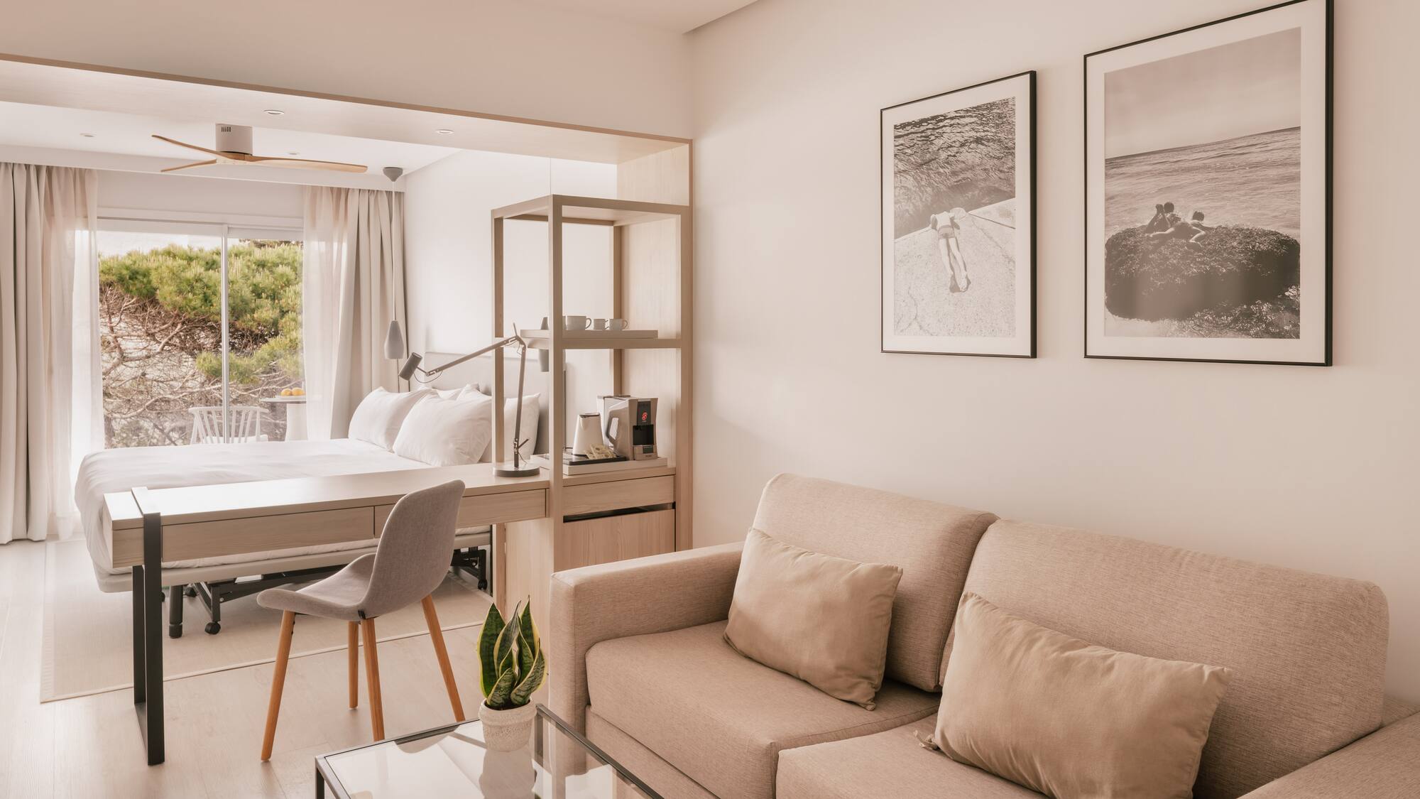 Modern hotel suite with beige sofa, desk, bed, and large window.