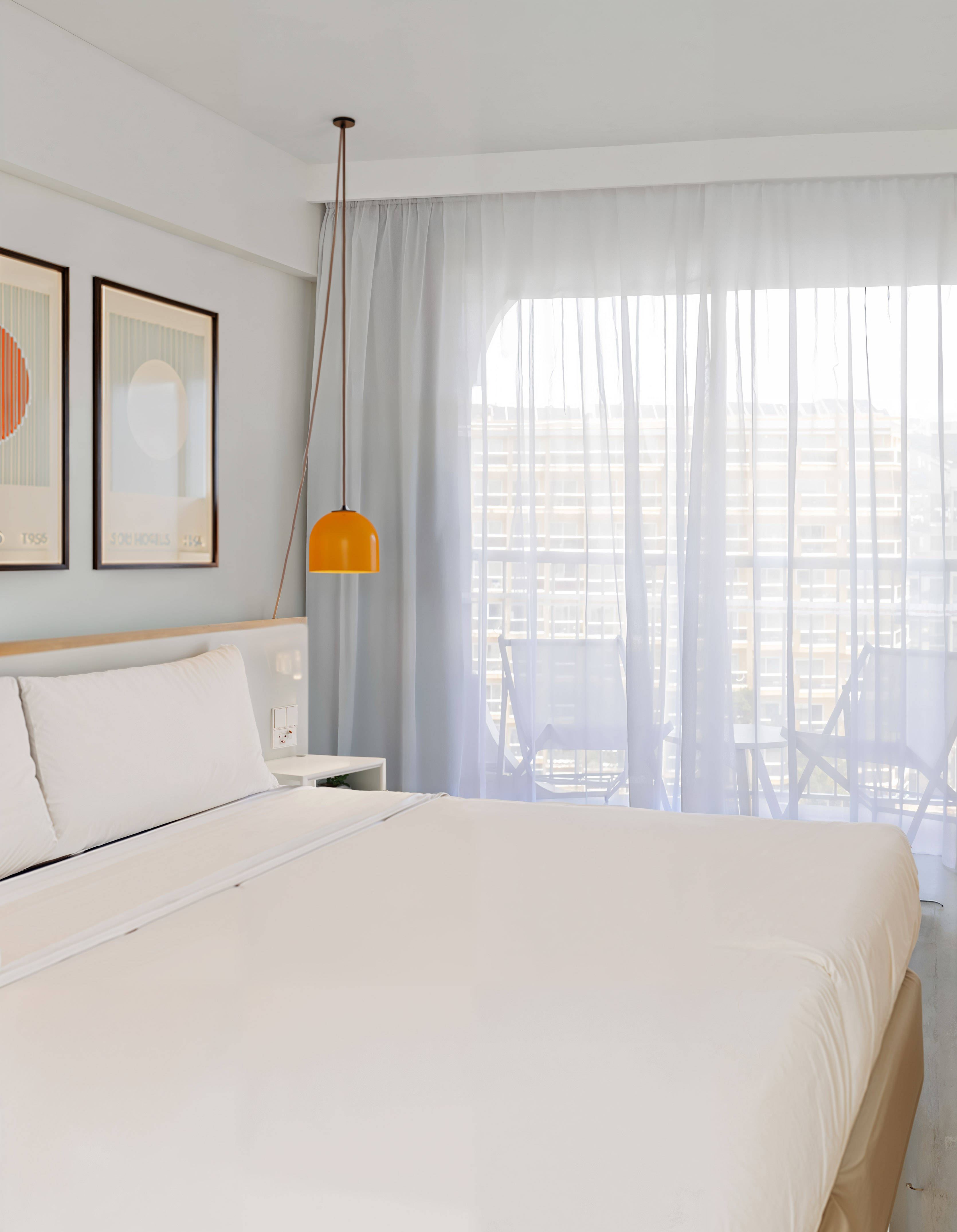 Modern hotel bedroom with white bed, orange pendant lamp, and balcony view.
