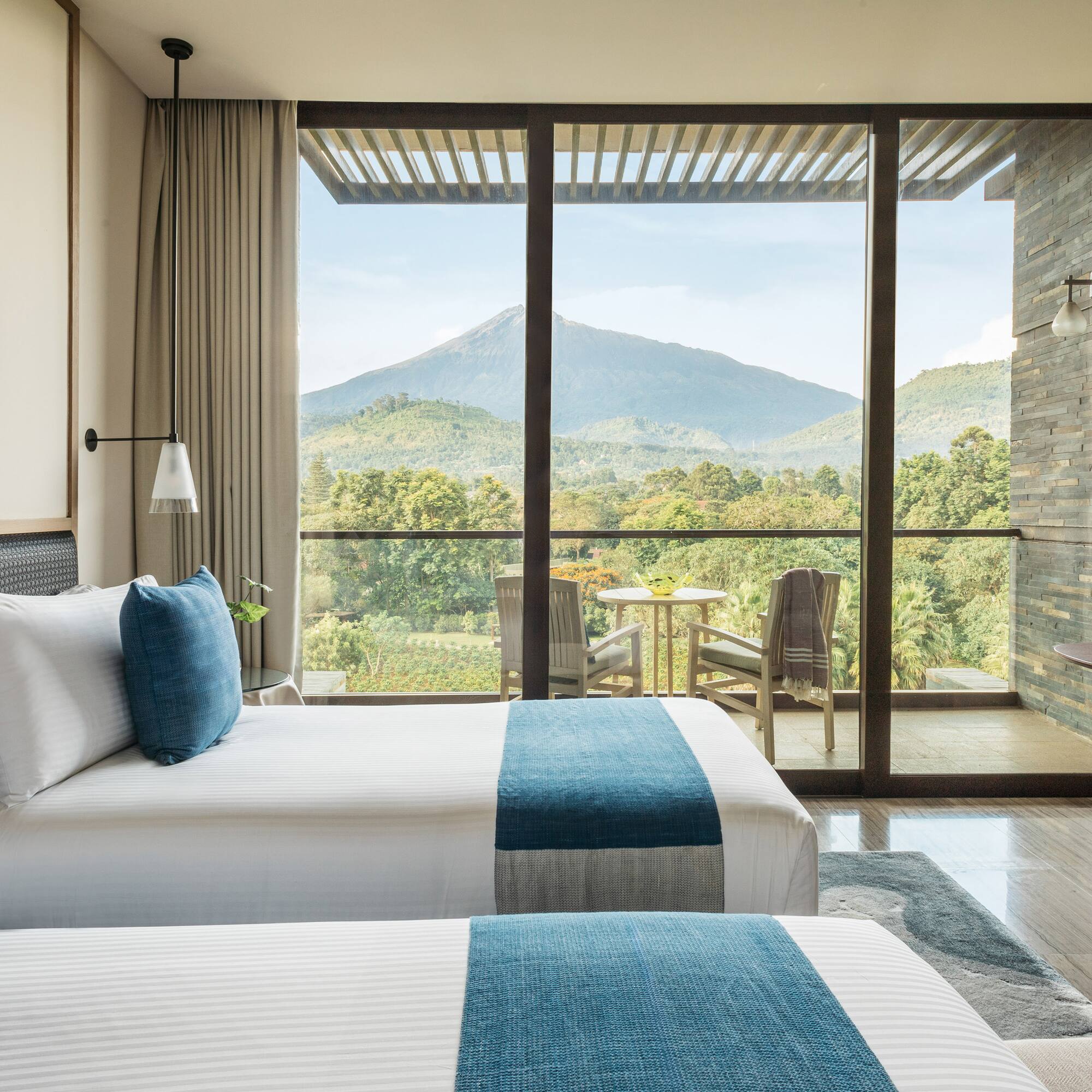 Modern hotel room with twin beds and balcony overlooking lush mountains.