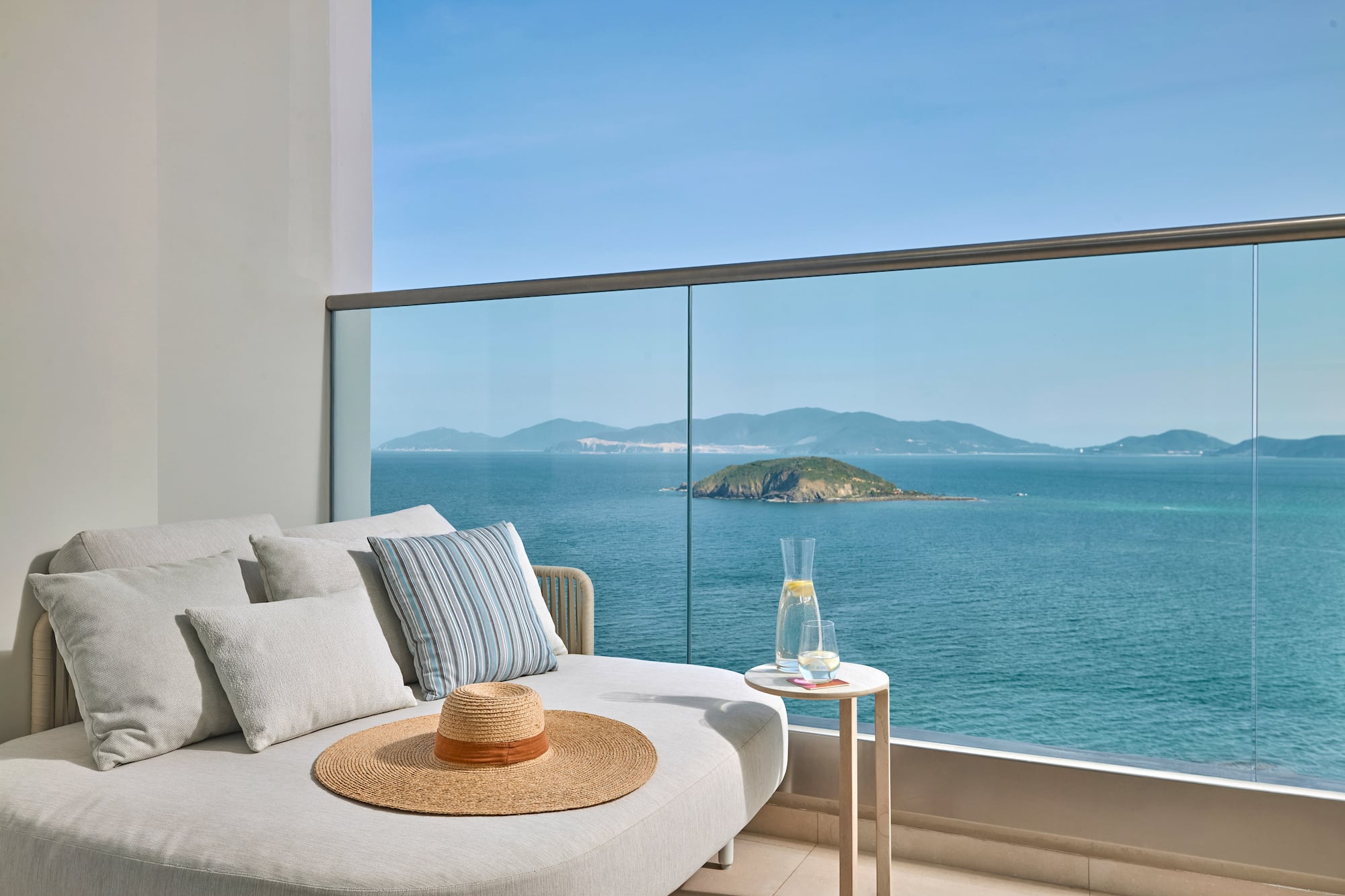Balcony daybed with ocean island view, straw hat, and refreshing drink.
