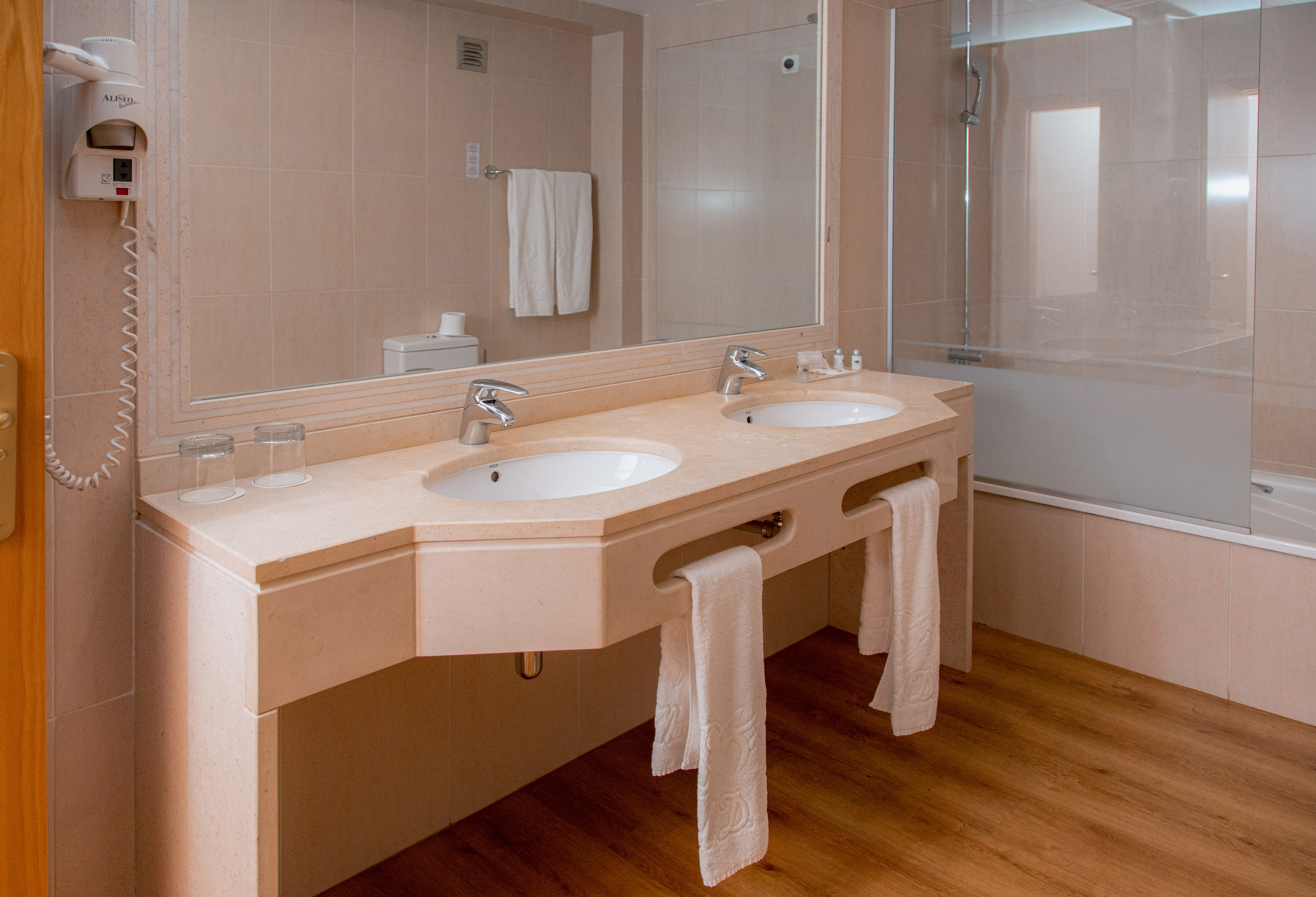 Modern hotel bathroom with double vanity sinks, mirror, bathtub, and wooden floor.