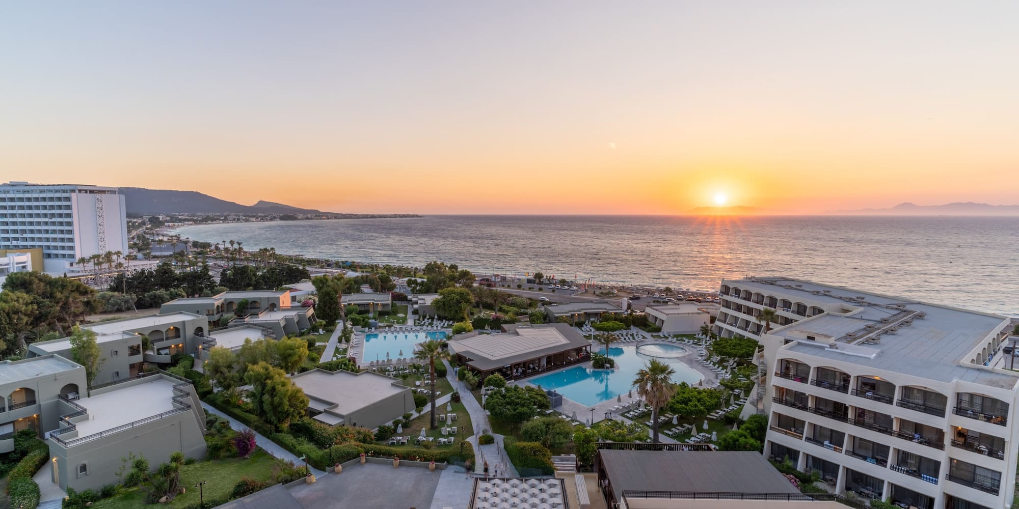 Aerial view of beachfront resort, multiple pools, and sunset ocean.