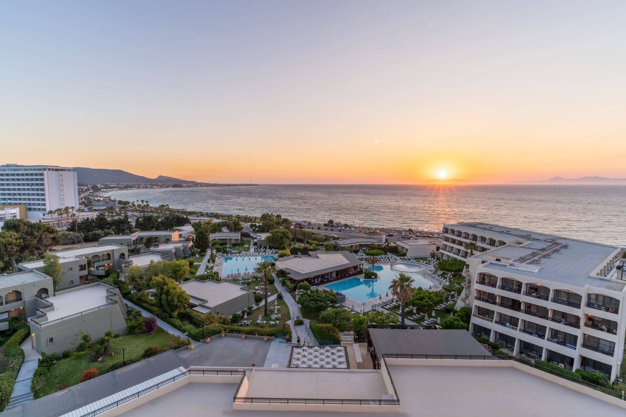 Aerial view of beachfront resort, multiple pools, and sunset ocean.