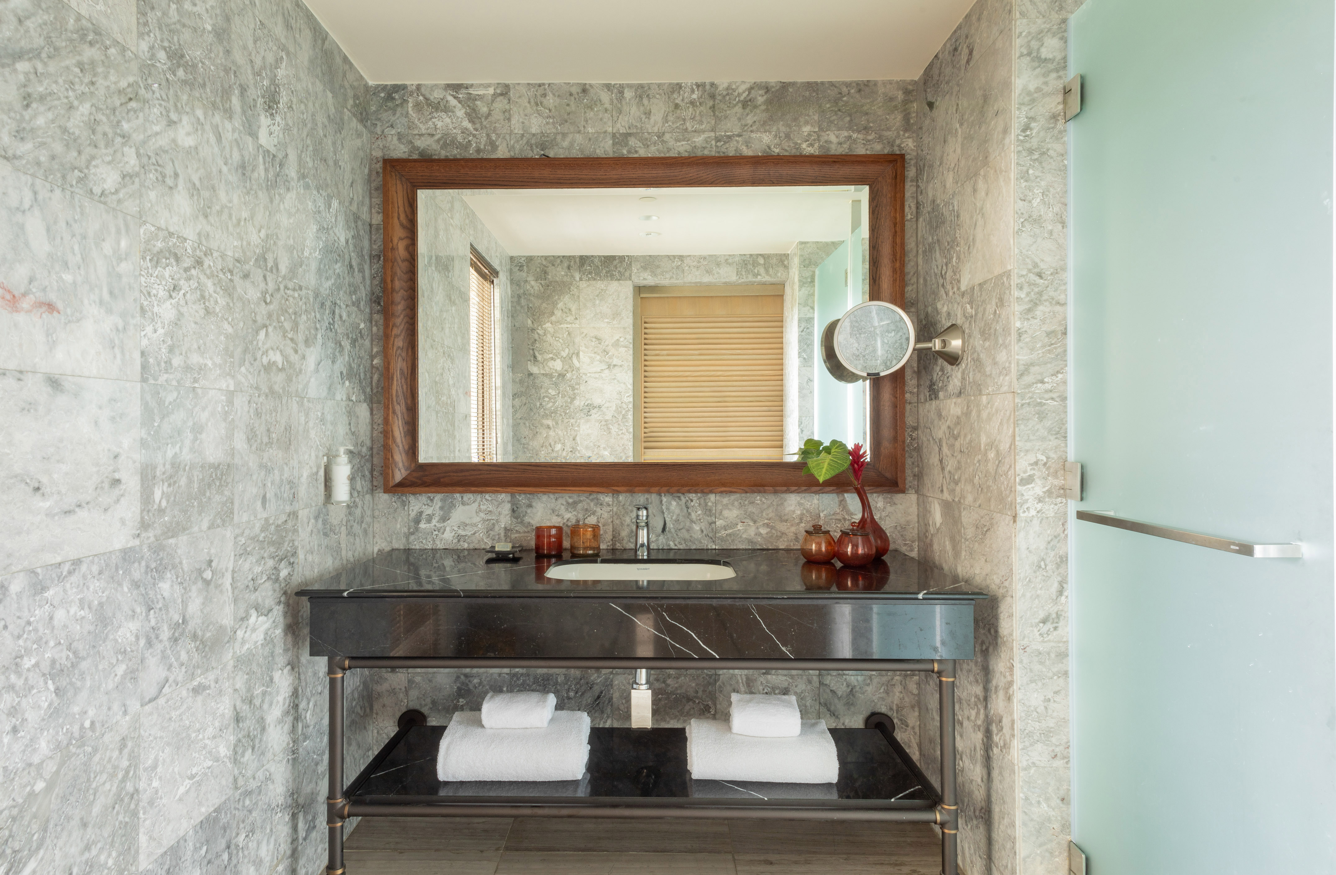 Modern hotel bathroom vanity with black marble counter, wood mirror, grey tiled walls.
