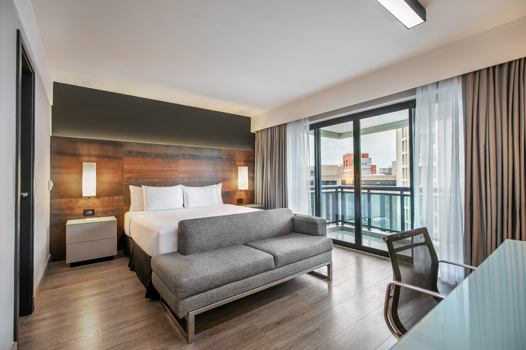 Quarto de hotel moderno com cama de casal, sofá cinza, escrivaninha e varanda. - Deluxe Queen Room com Varanda - Meliá Ibirapuera, São Paulo