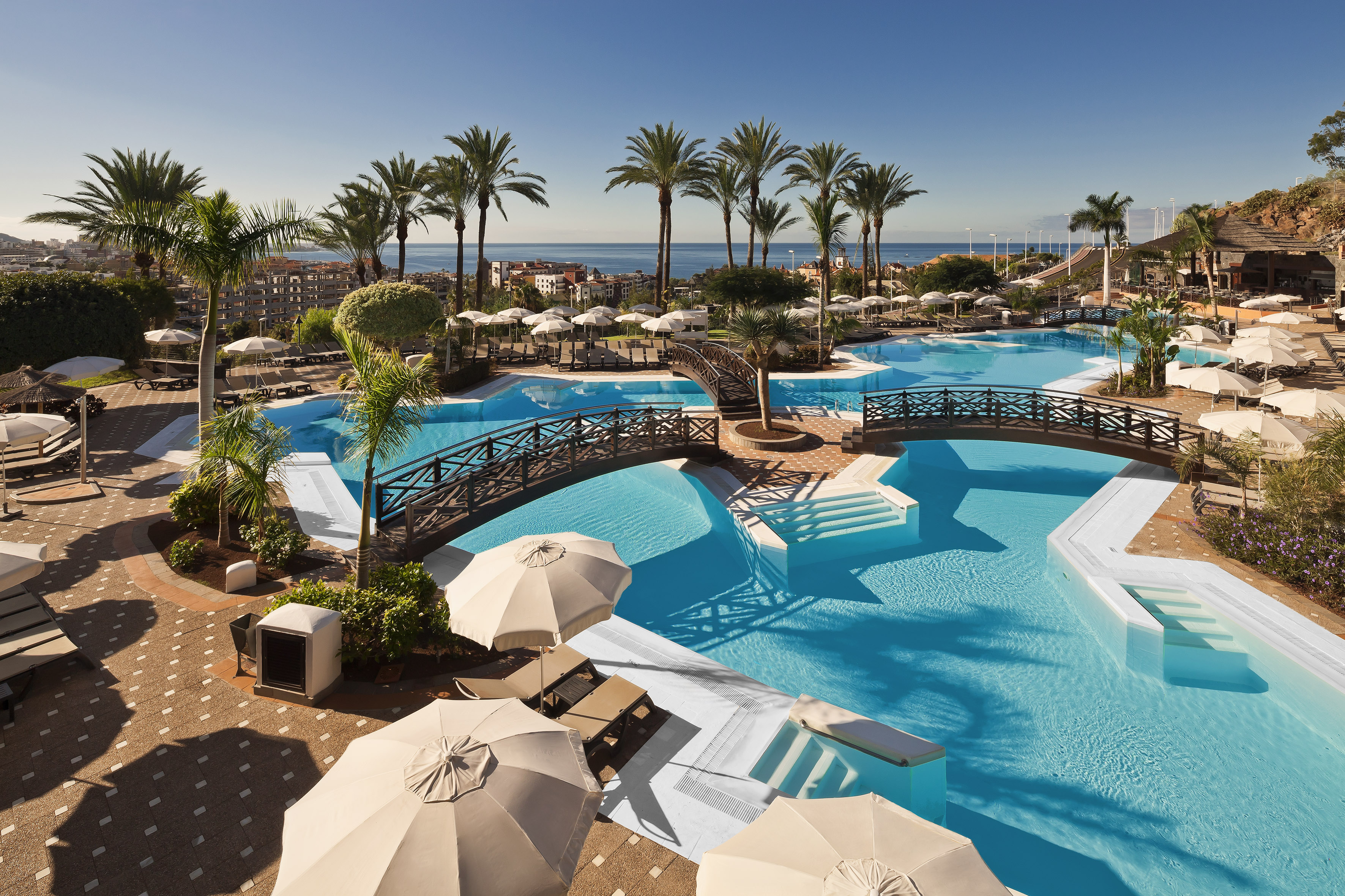 Sunny resort pool complex with bridges, palm trees, and ocean view.