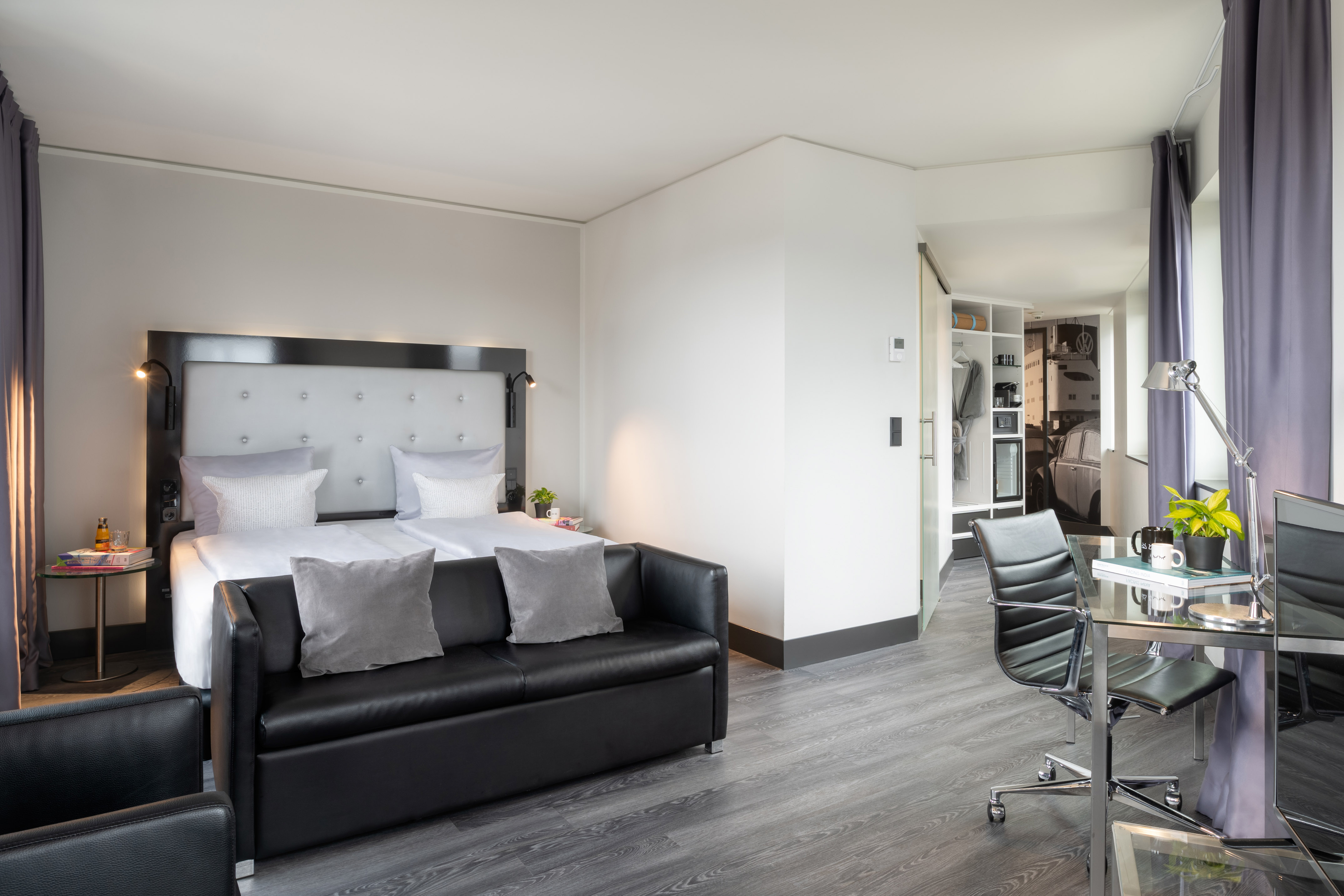 Modern hotel room with queen bed, black sofa, glass desk, open closet.