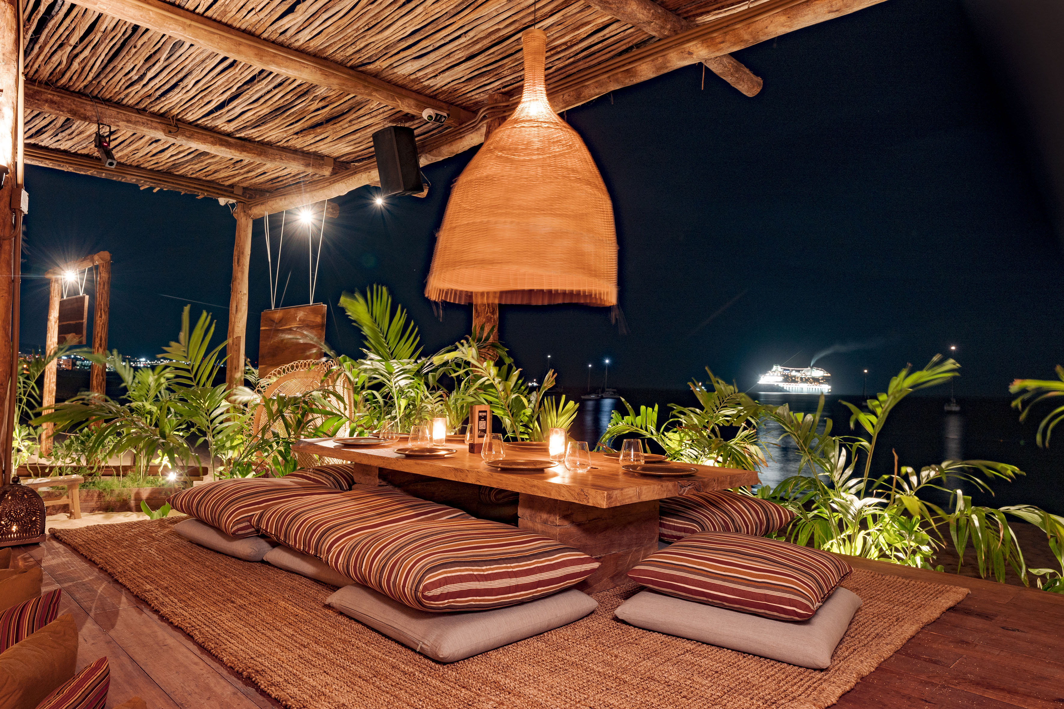 Tropical outdoor dining at night with wooden table, floor cushions, and ocean view.