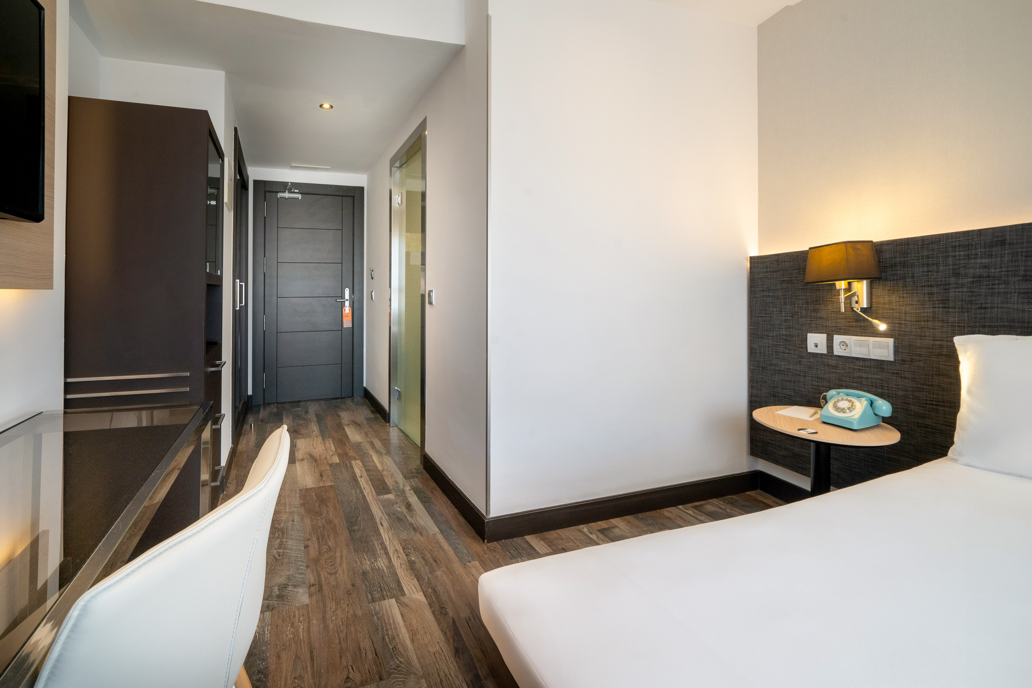 Modern hotel room with white bed, desk, dark wardrobe, and wooden floor.