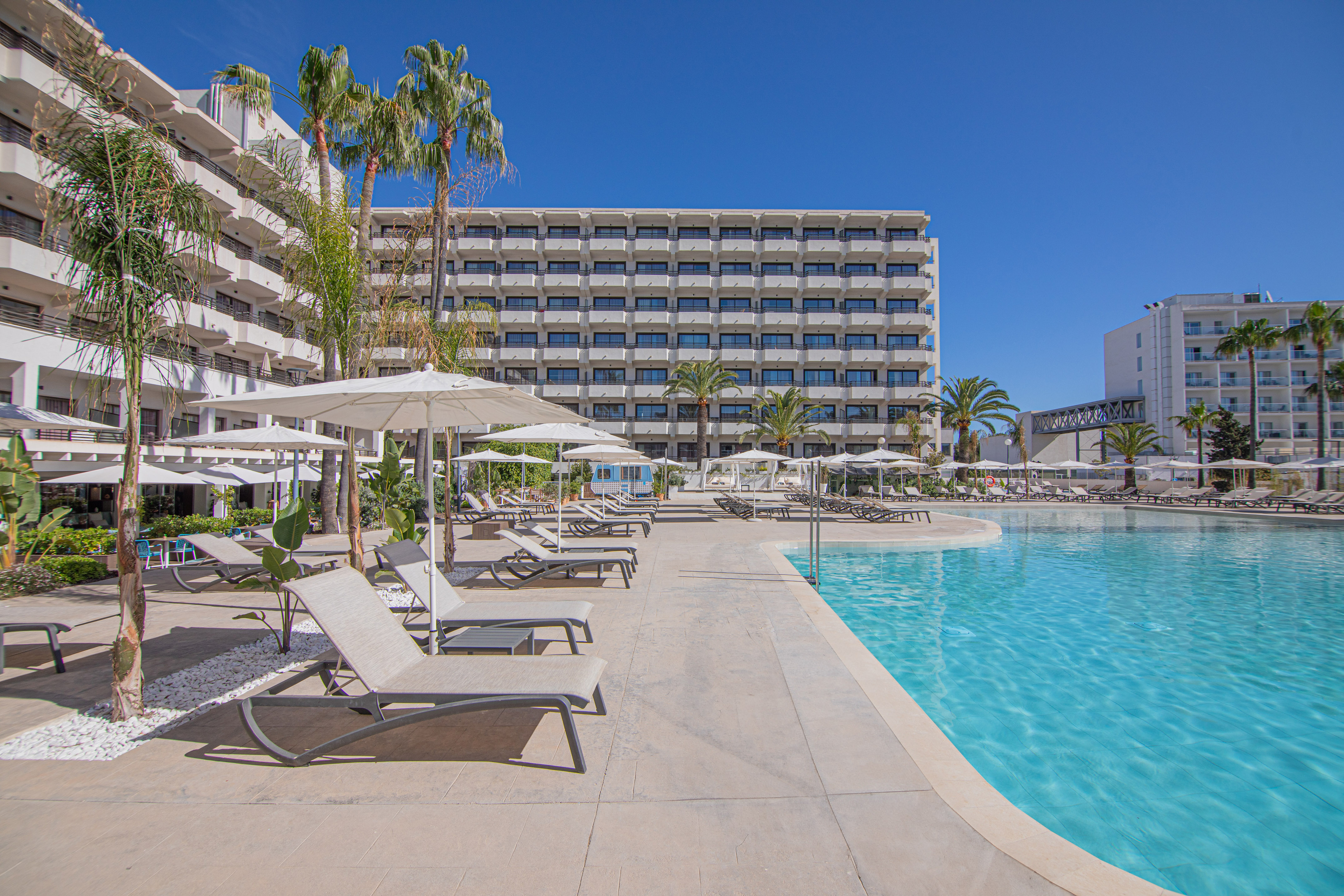 Tropical resort pool deck with sun loungers, palm trees, and hotel buildings.