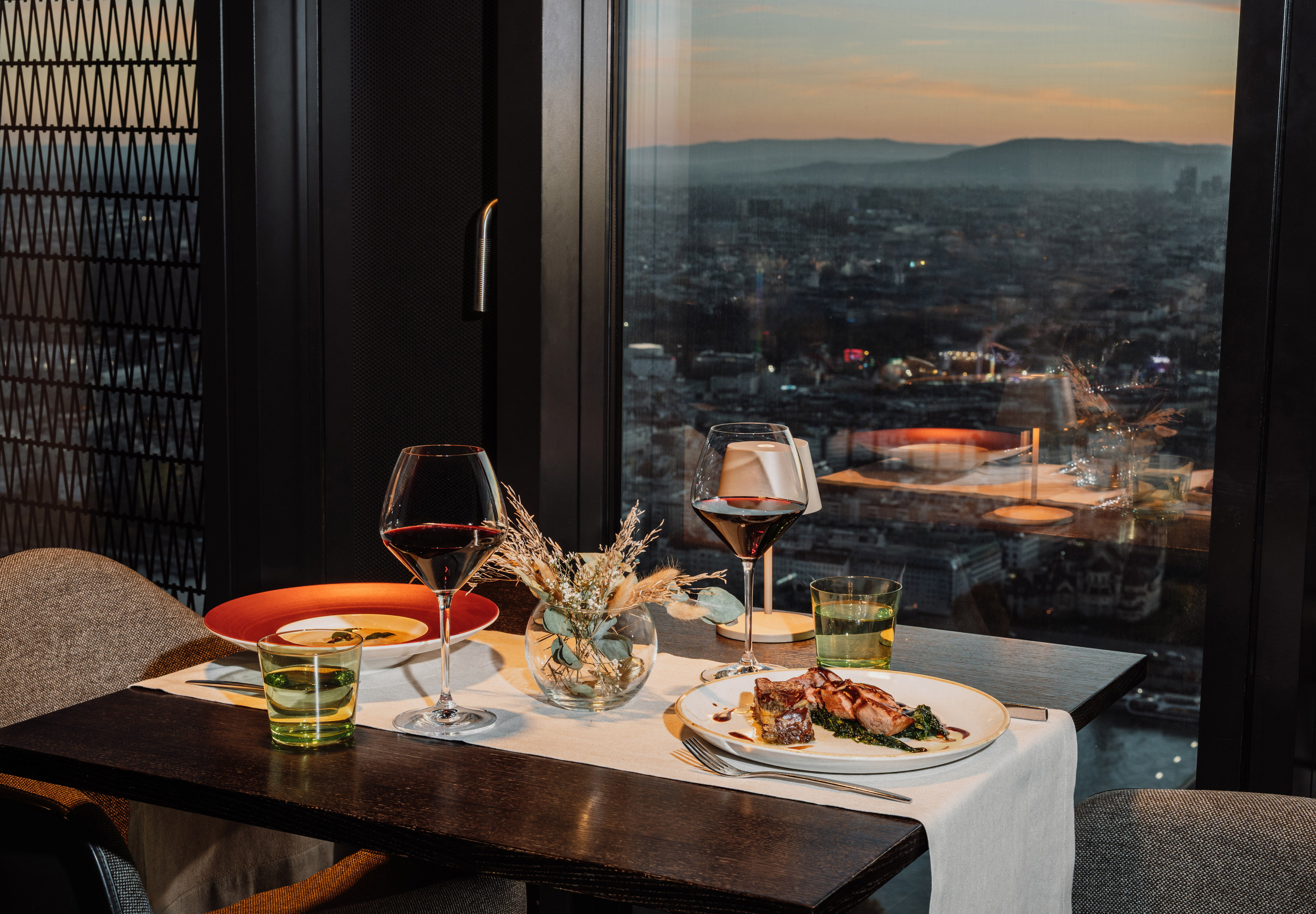 Fine dining dinner with red wine overlooking city skyline at dusk.
