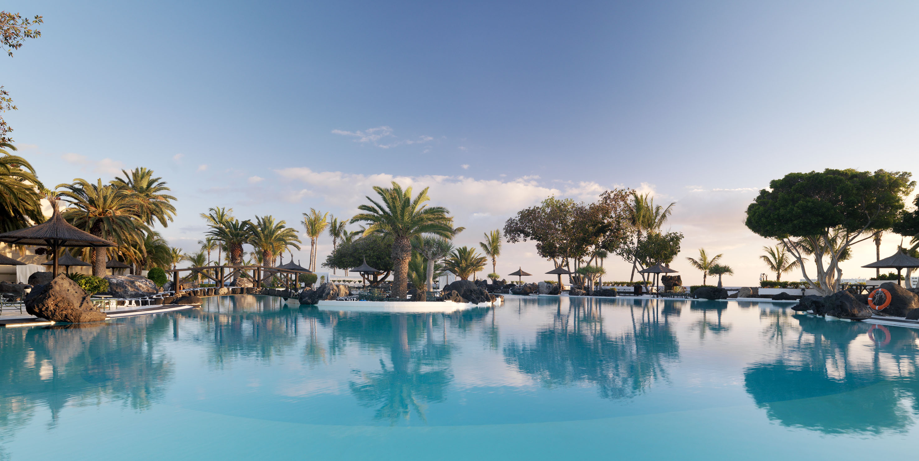 Tropical resort swimming pool with palm trees, cabanas, and blue sky.