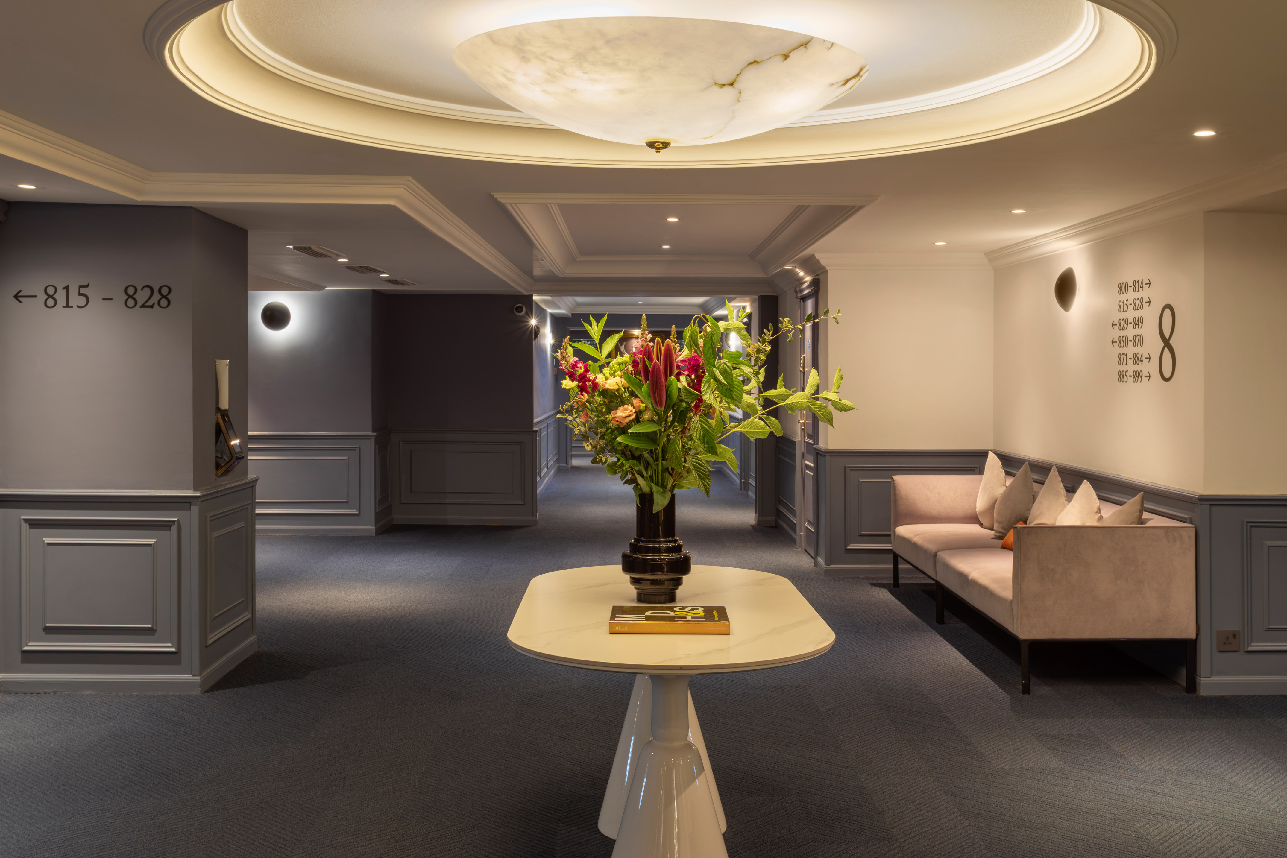 Elegant hotel corridor with floral arrangement, sofa, and room number signs.