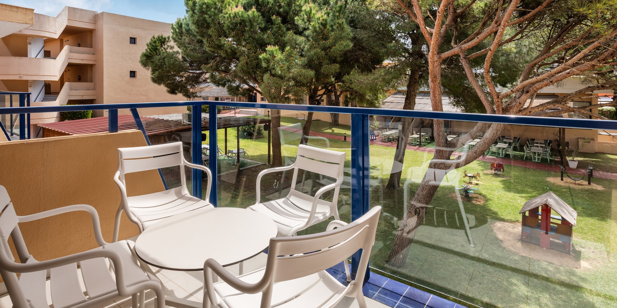 Hotel balcony with white patio furniture, garden, trees, and playground.