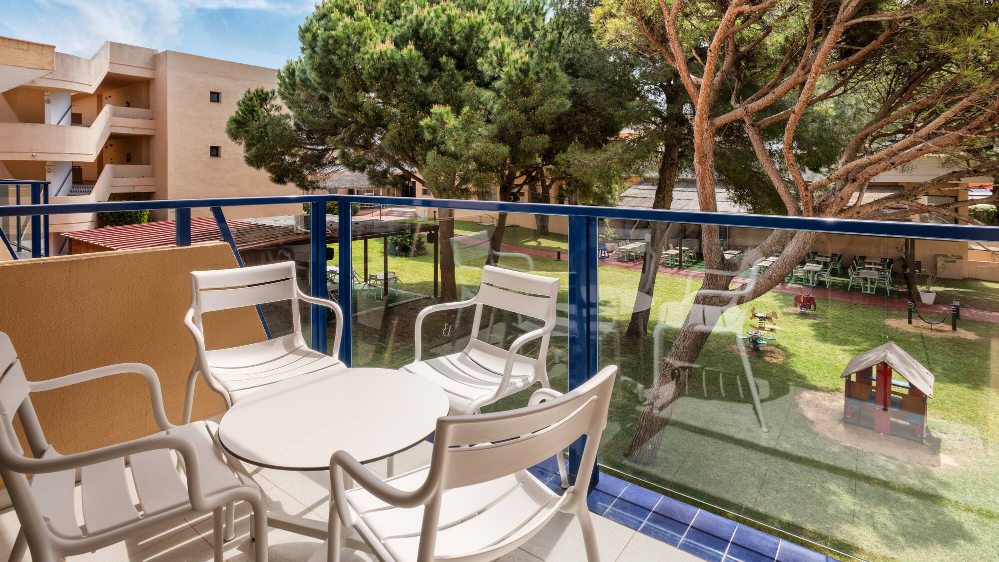 Resort balcony with white patio set, view of garden, playground, and large trees.