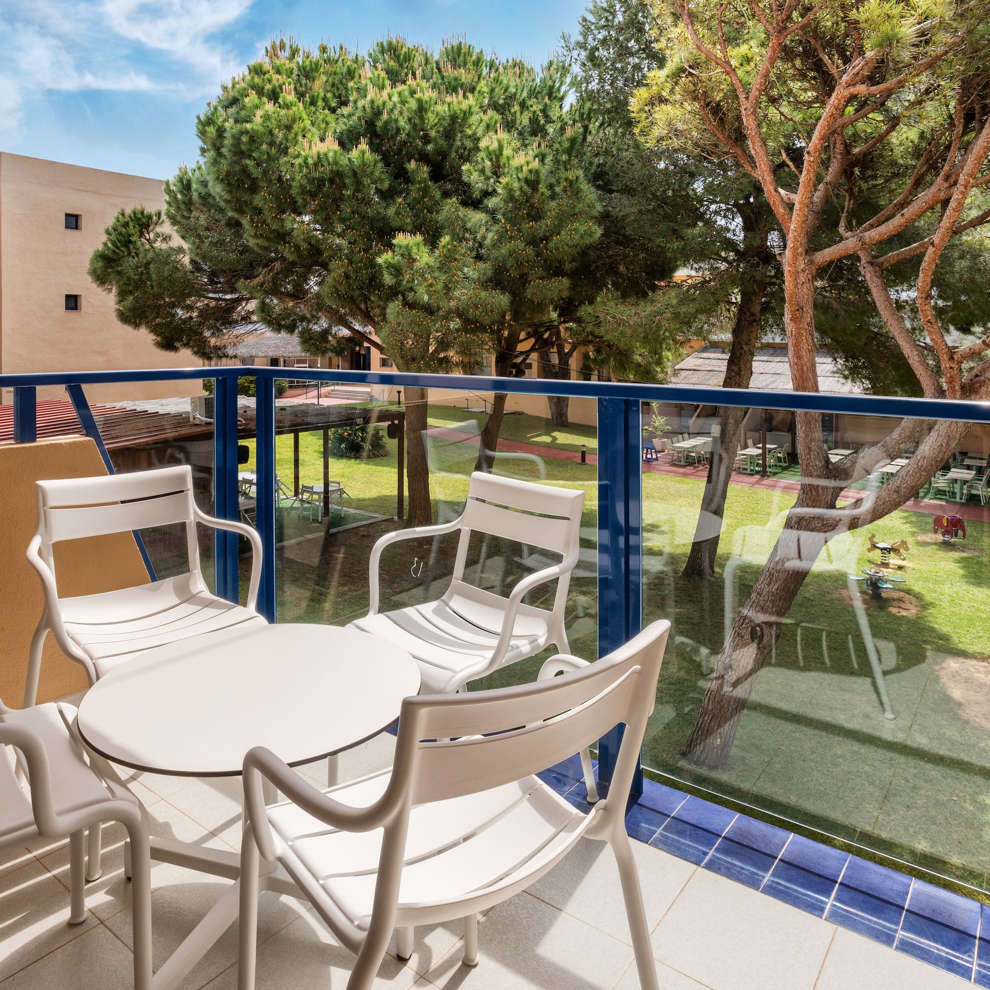 Hotel balcony with white patio furniture, garden, trees, and playground.