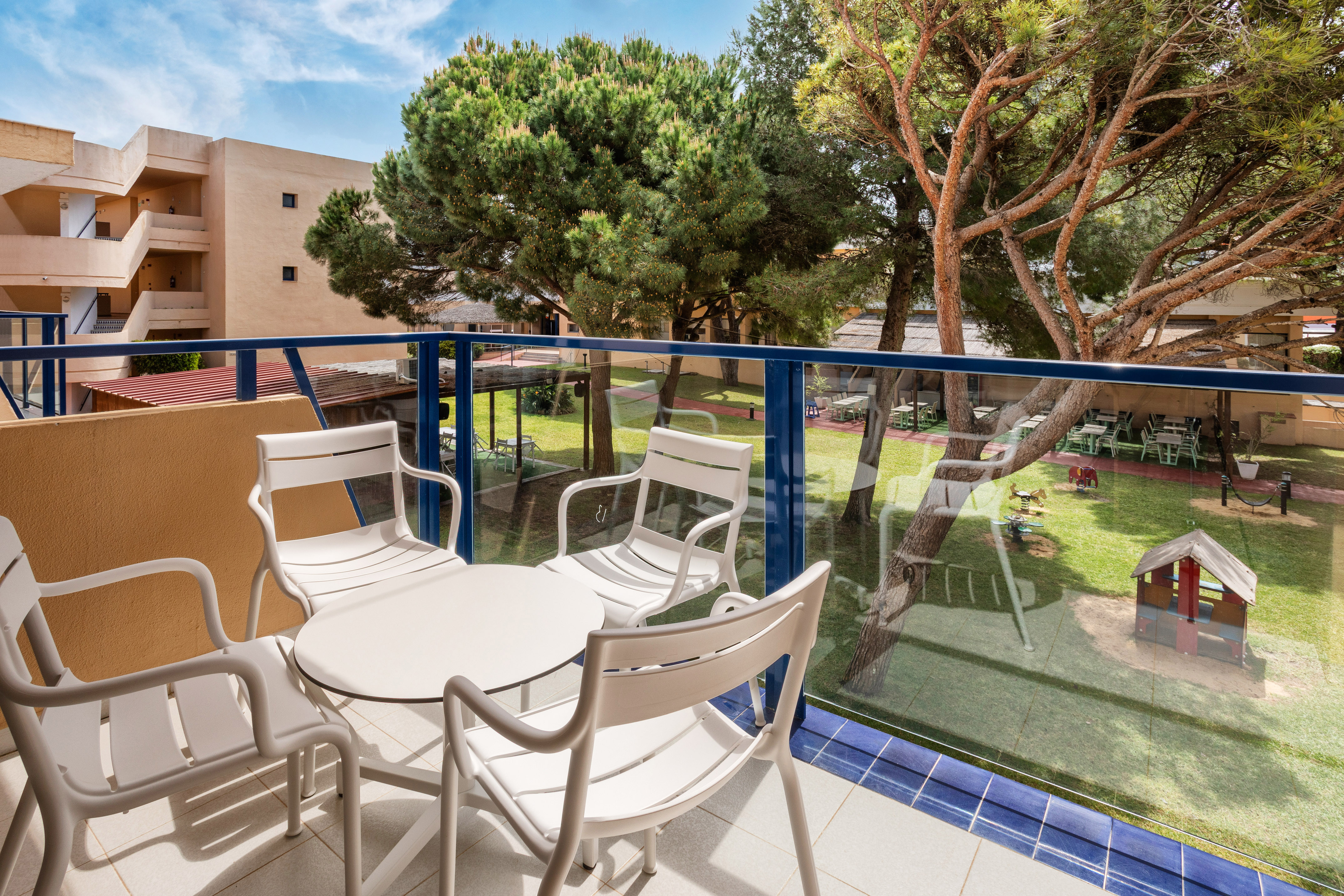 Resort balcony with white patio set, view of garden, playground, and large trees.