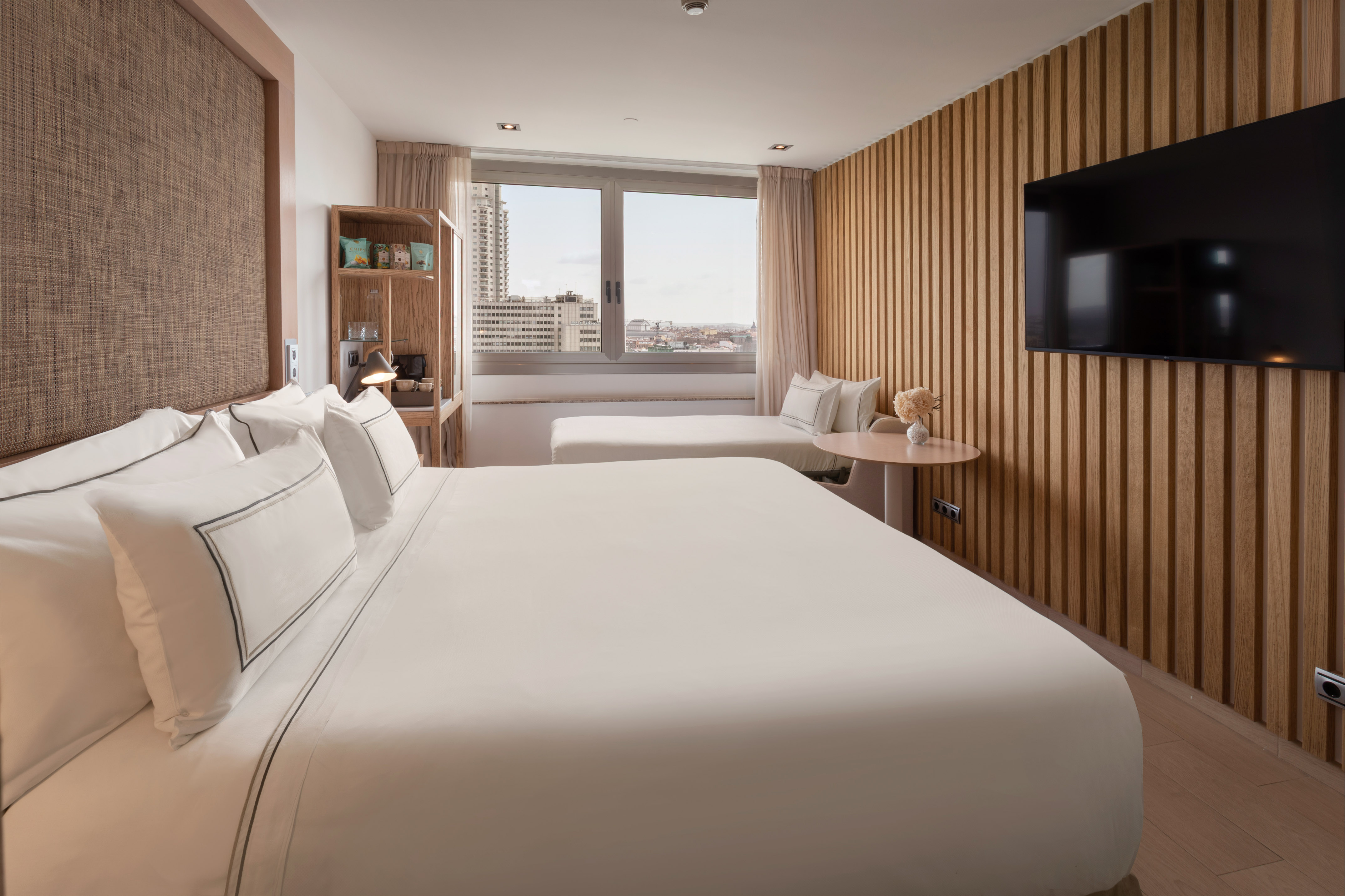 Modern hotel bedroom with city view, white bed, wooden wall, and mounted TV.