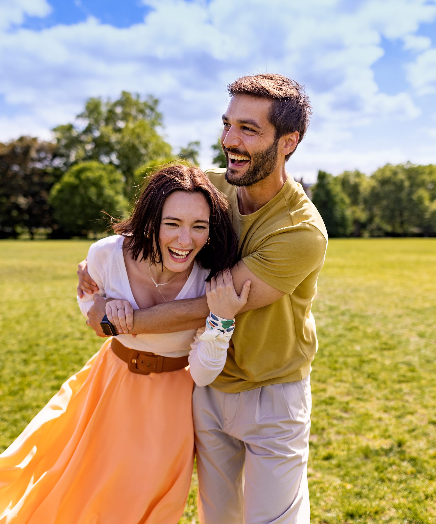 Joyful couple embracing and laughing in a vibrant green park on a sunny day.