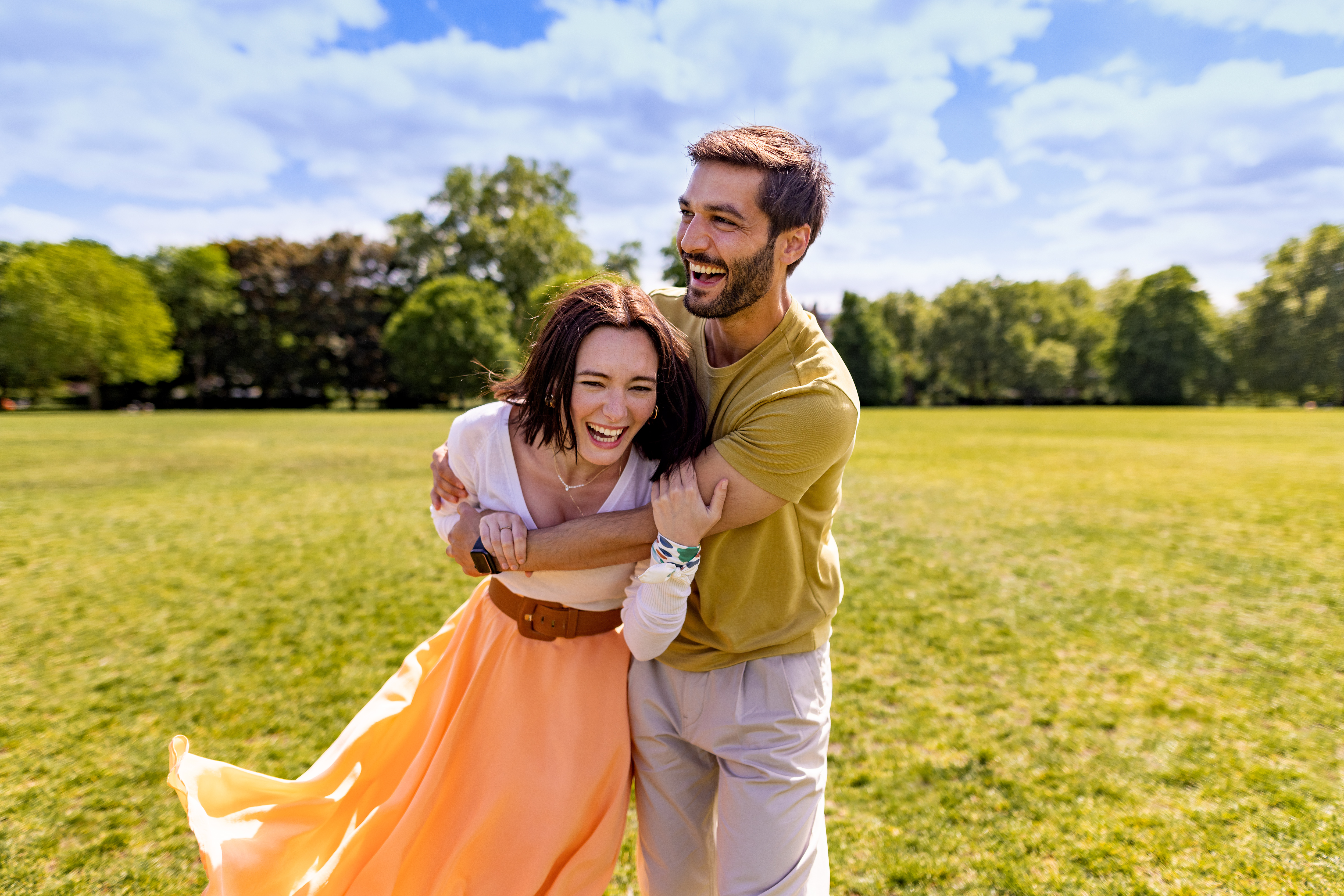 Joyful couple embracing and laughing in a vibrant green park on a sunny day.