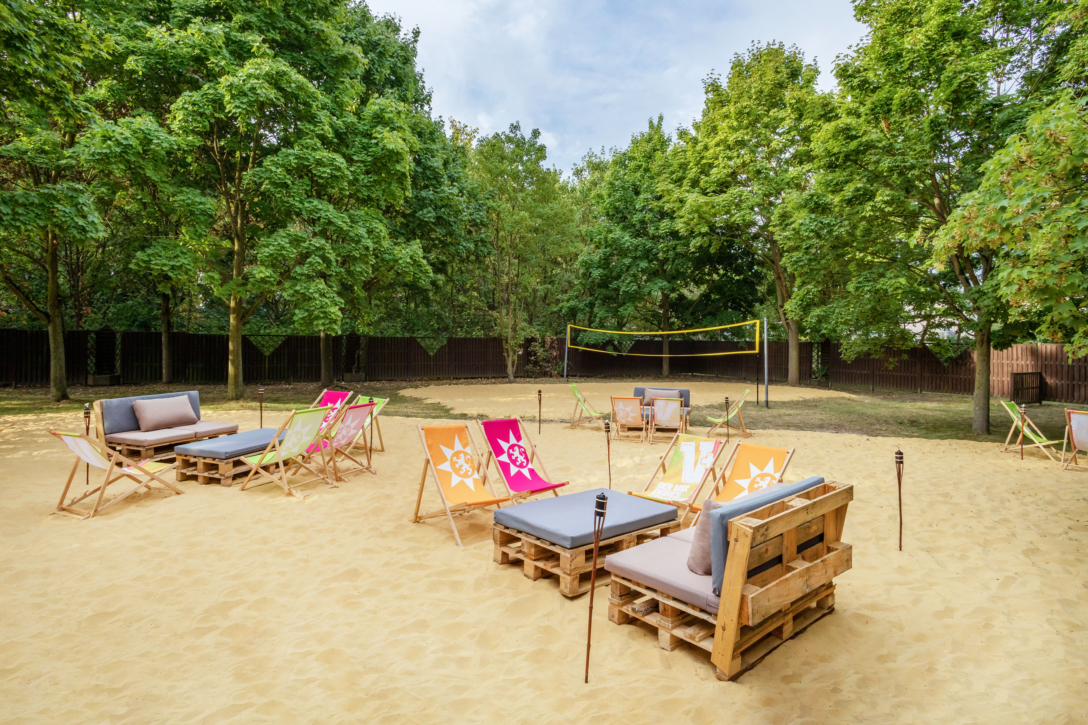 Outdoor sand lounge with pallet sofas, colorful chairs, and volleyball net.
