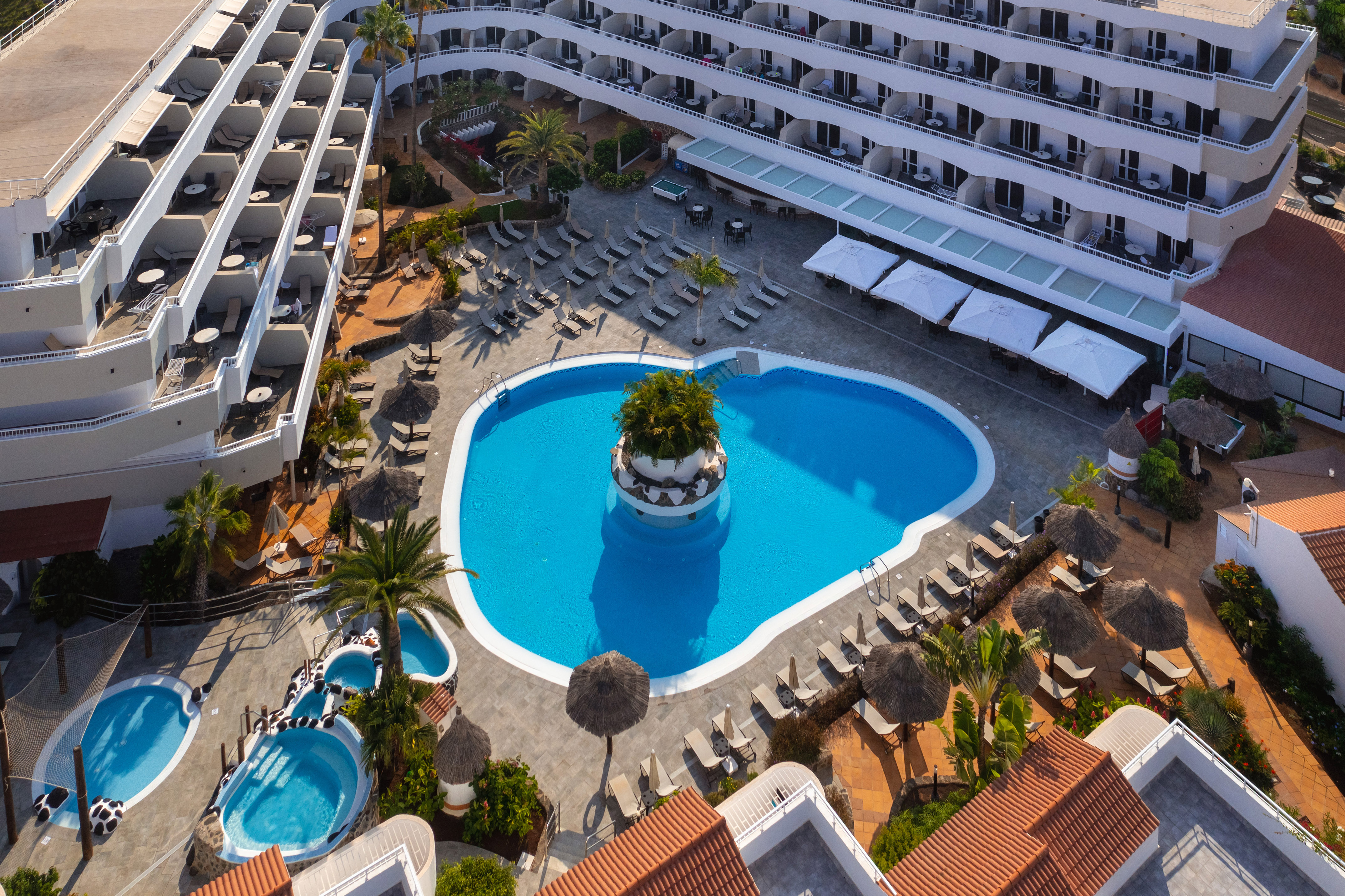 Aerial view of resort hotel with large blue swimming pool, loungers, and palm trees.