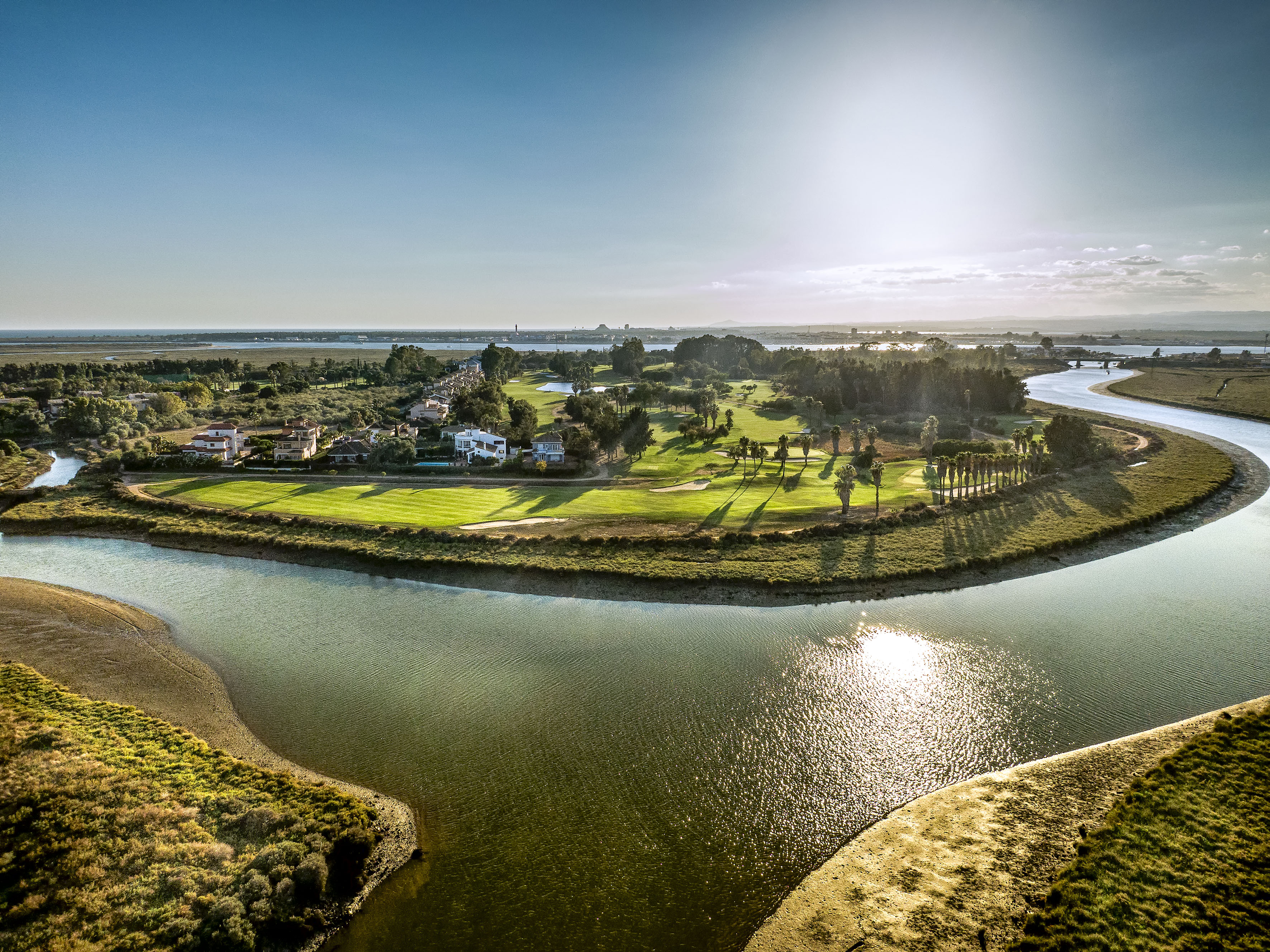 Sunny aerial view of a riverside golf course with homes and palm trees.