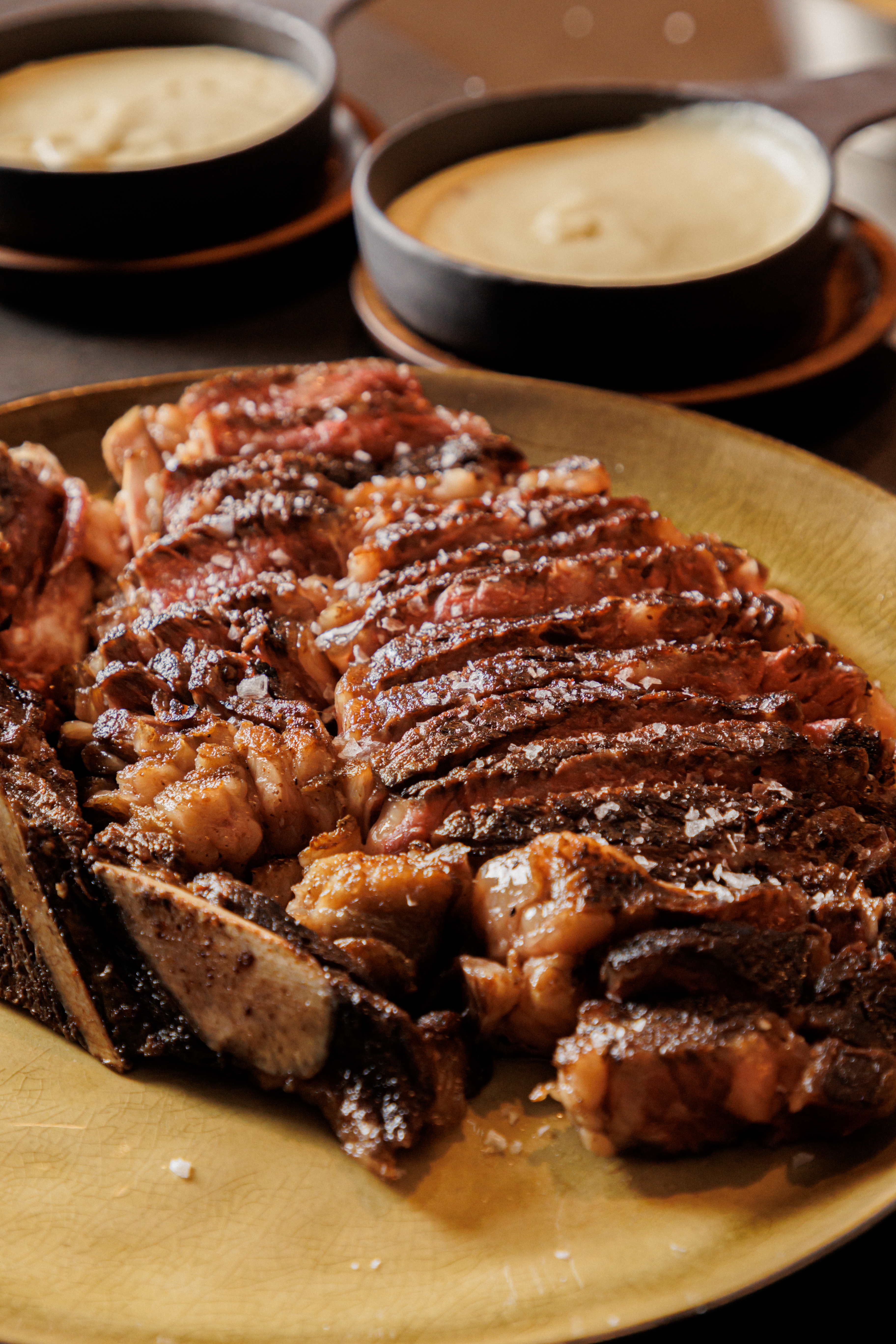 Sliced grilled steak with bone, flaked salt, and creamy sauces.