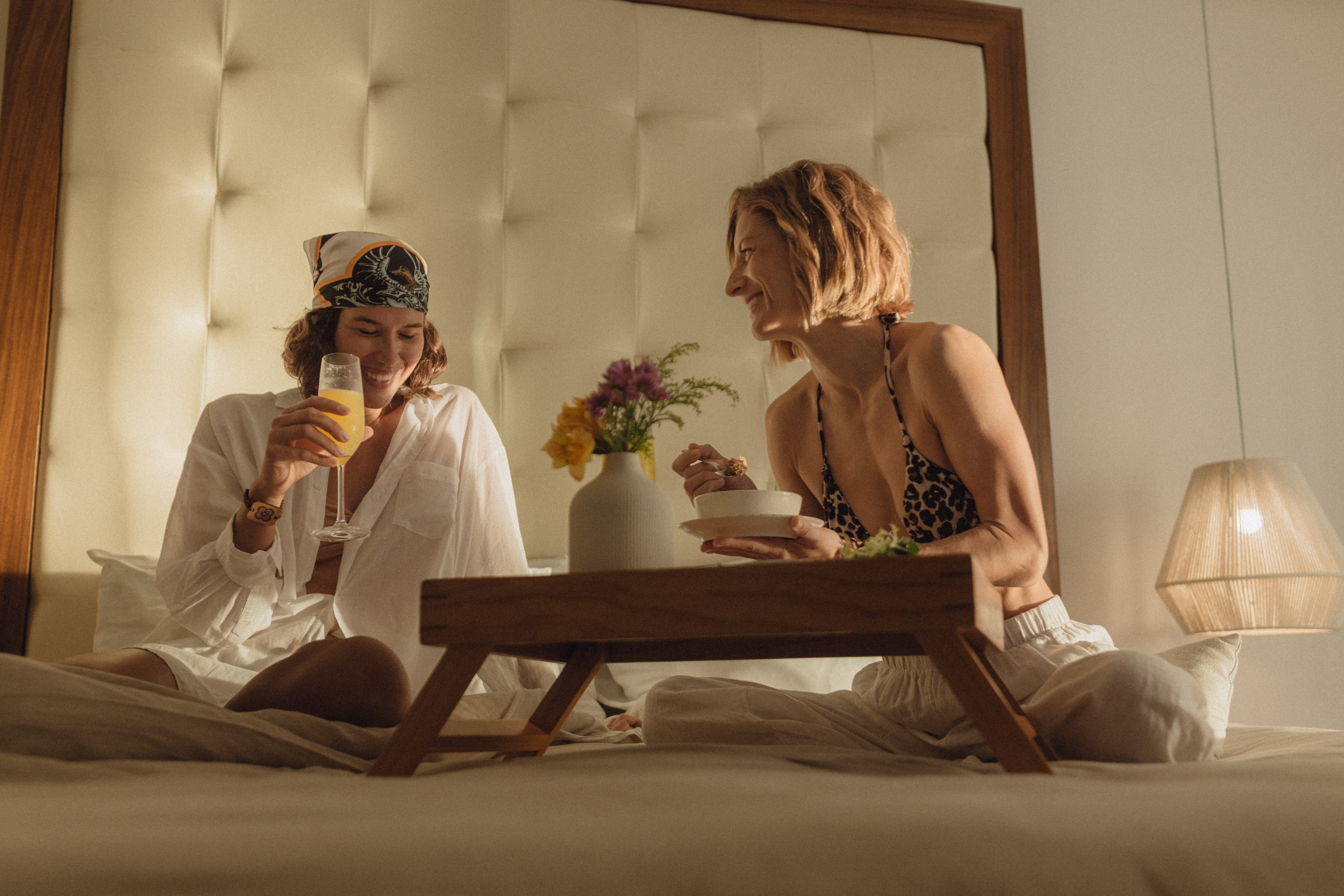 Smiling women enjoying breakfast in bed with juice and flowers.