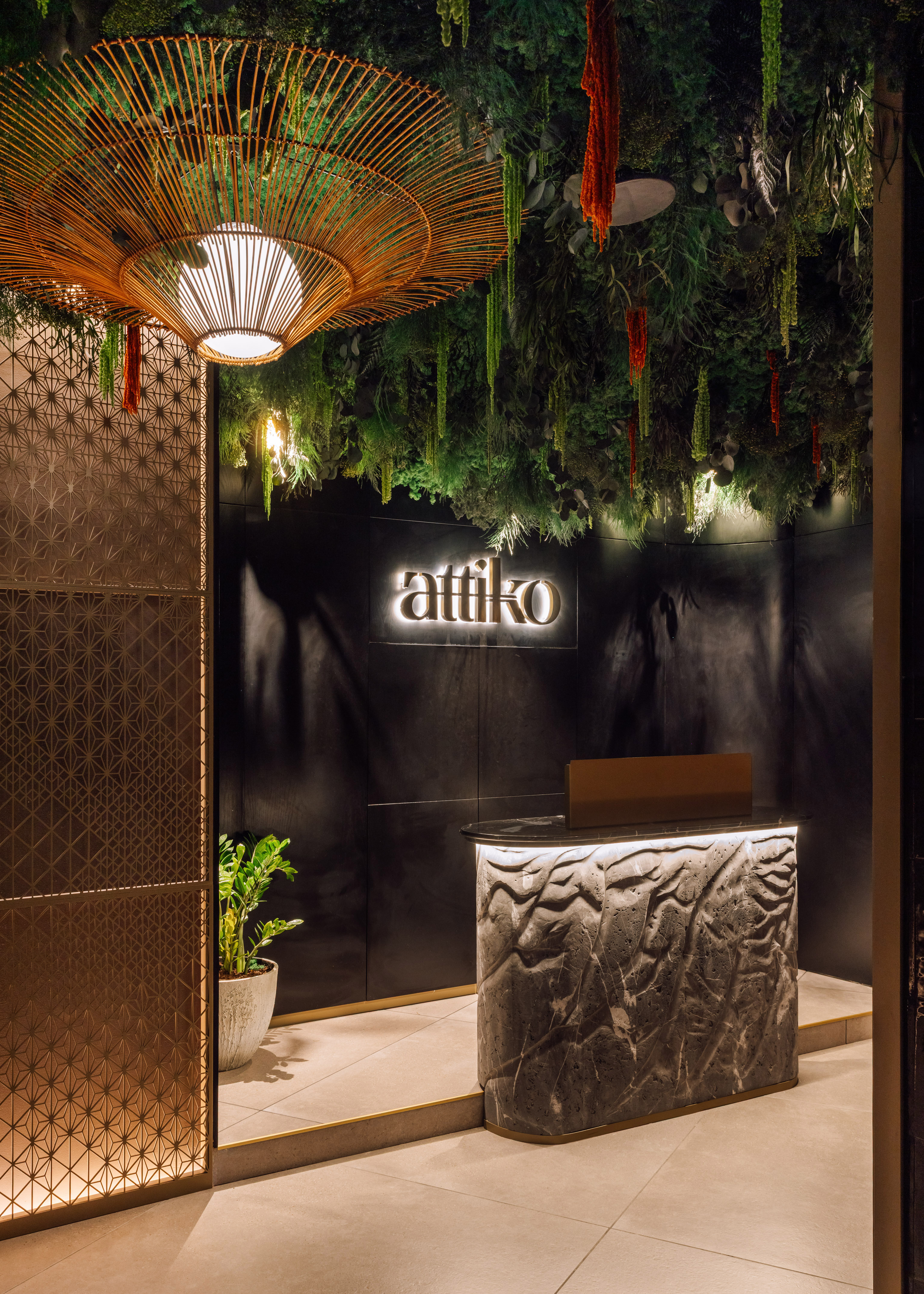 Attiko lounge reception: lush green ceiling, rattan chandelier, illuminated stone desk.