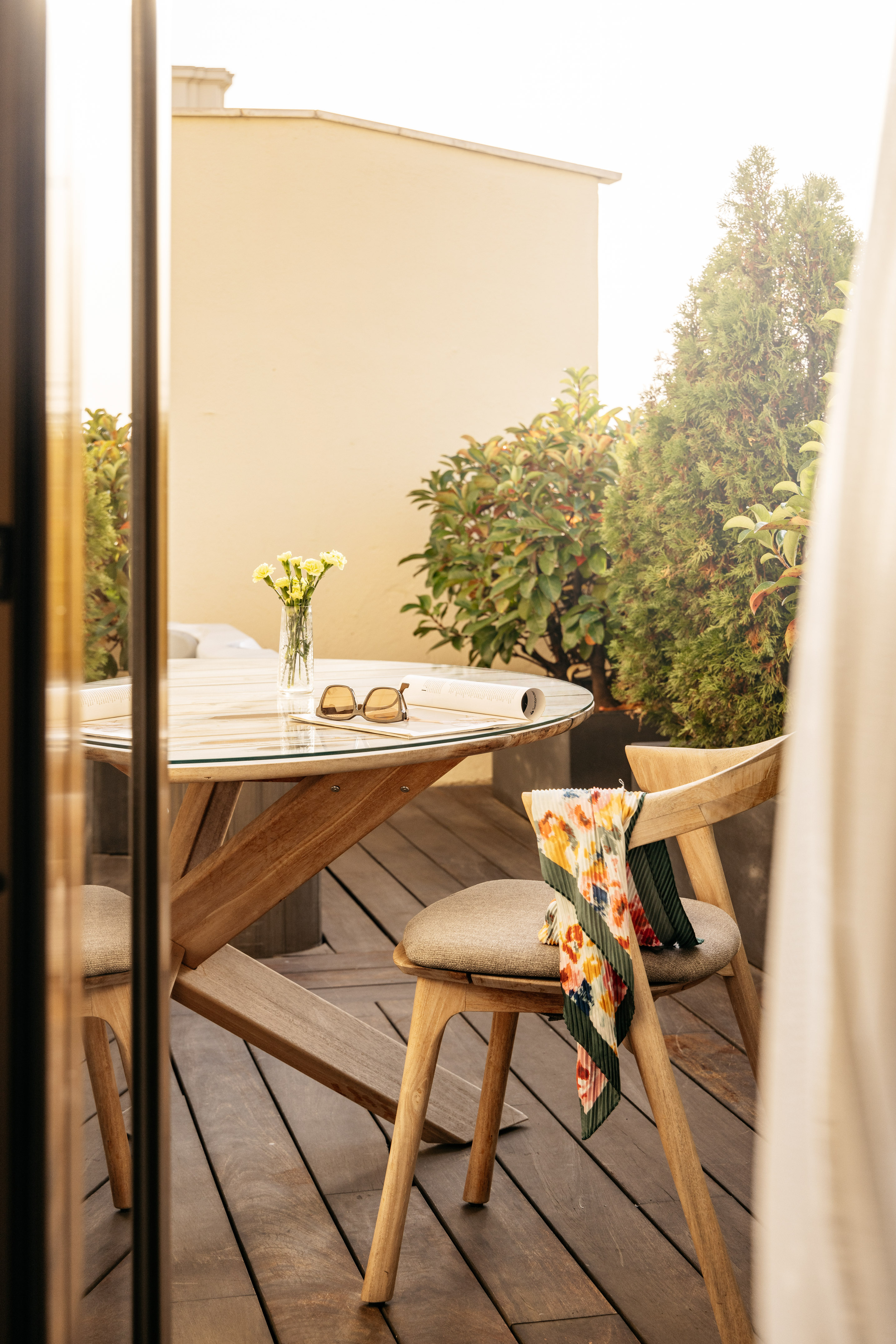 Sunny patio with wooden table, chairs, flowers, sunglasses, and colorful scarf.