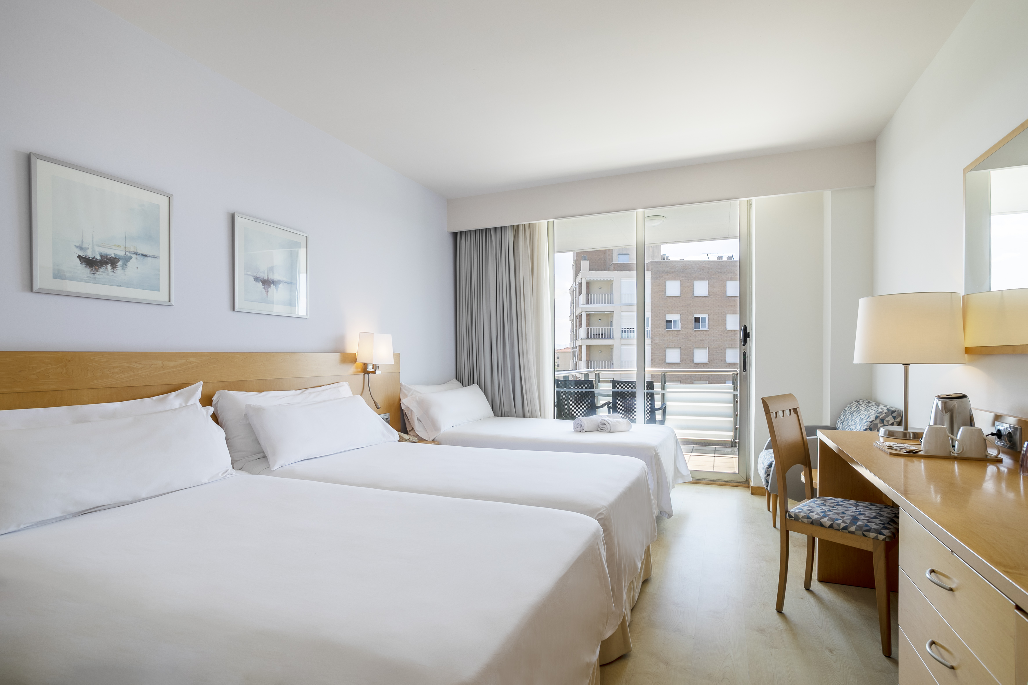 Bright triple hotel room with white beds, balcony, city view, and work desk.