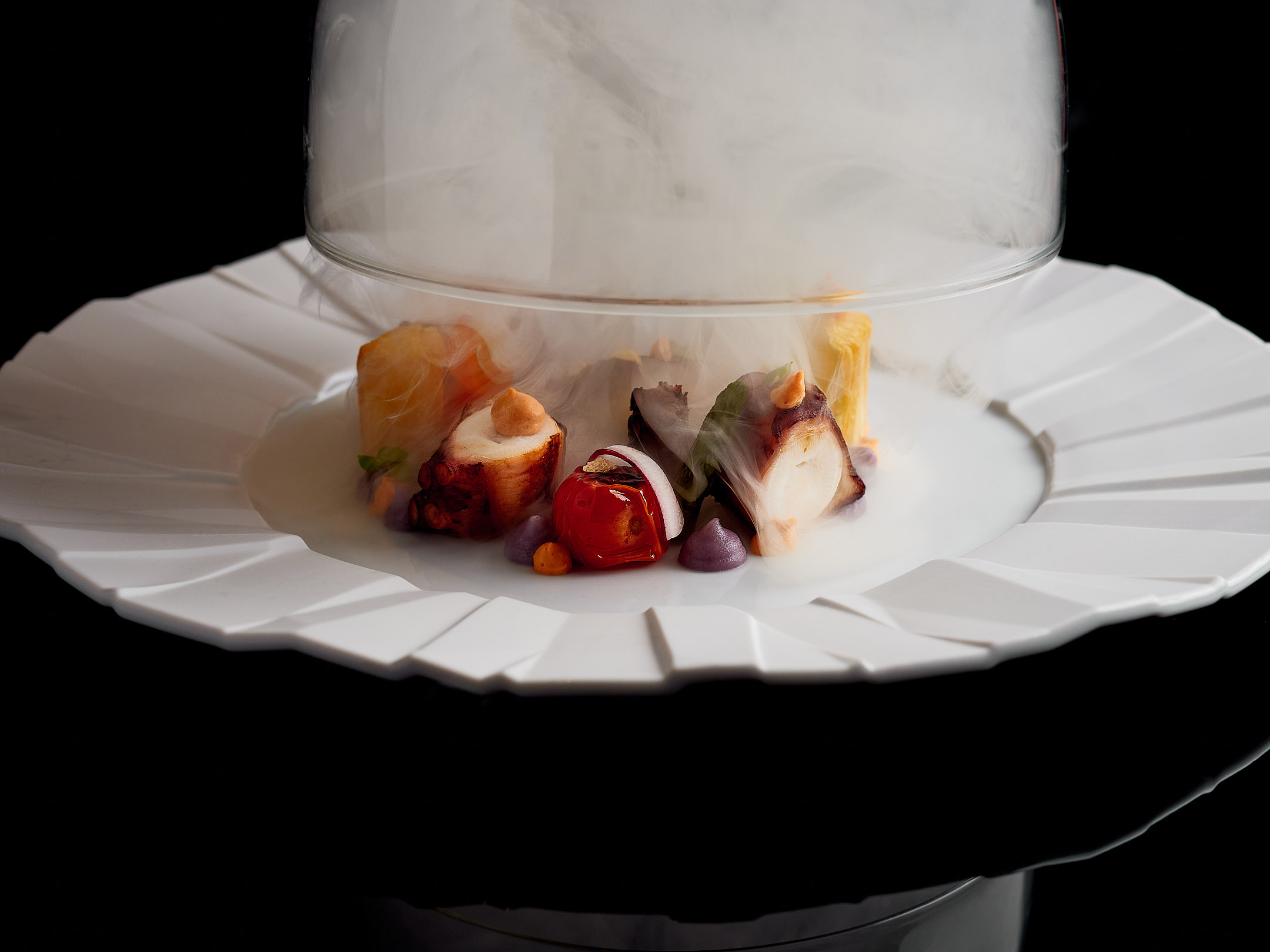 Fine dining smoked octopus under glass cloche on white geometric plate.