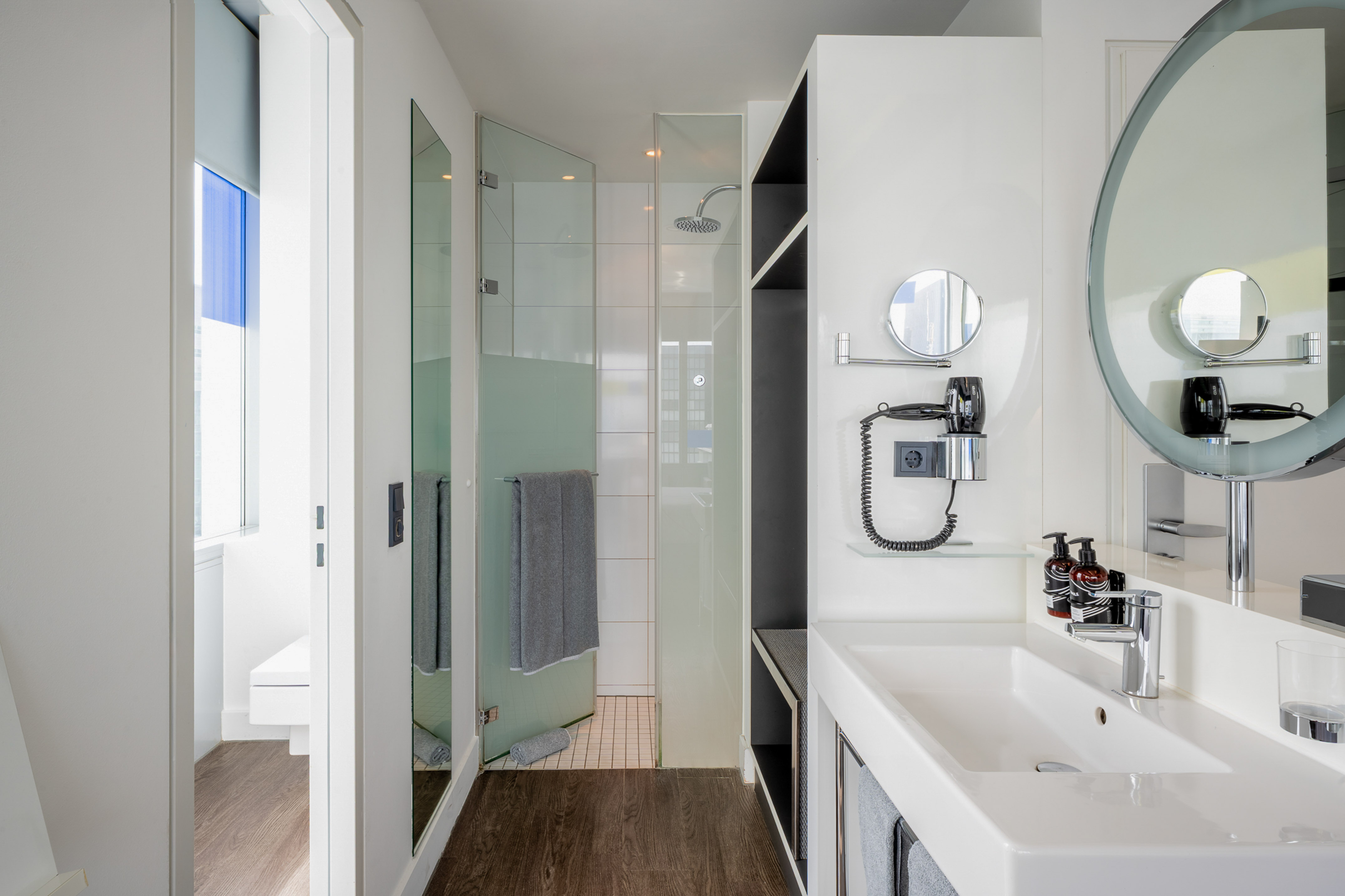 Modern hotel bathroom with white vanity, round mirror, walk-in shower, and dark wood floor.